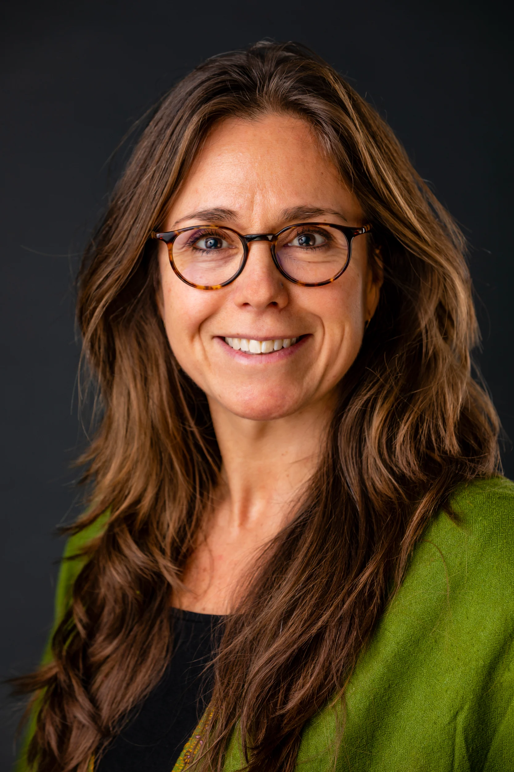 Portrætfoto af smilende kvinde med brunt hår, briller og grøn trøje mod mørk baggrund. Venligt udtryk.