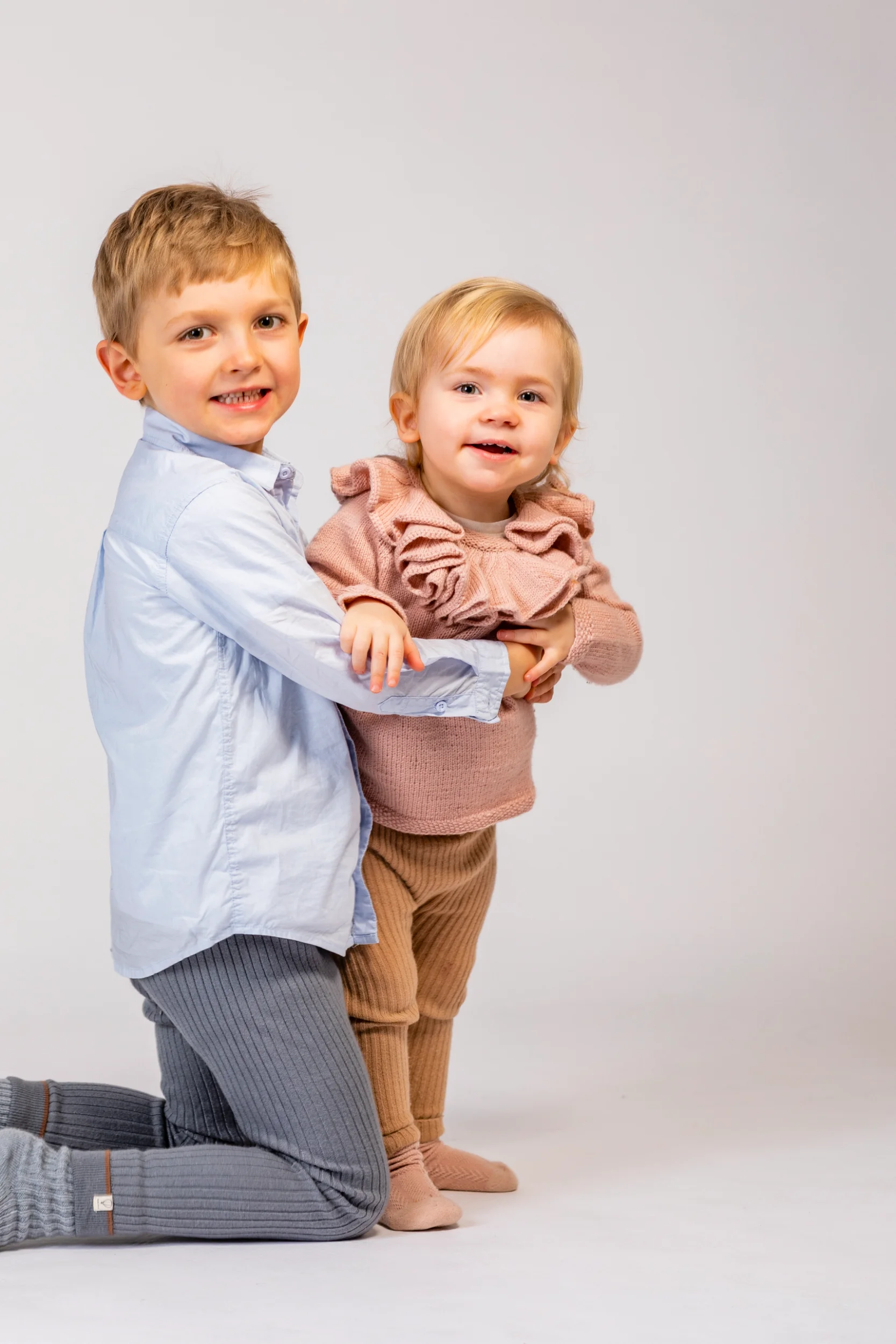 Børnefotografering af storebror på knæ, som holder sin lillesøster, der smiler bredt mod kameraet i et hvidt studie.