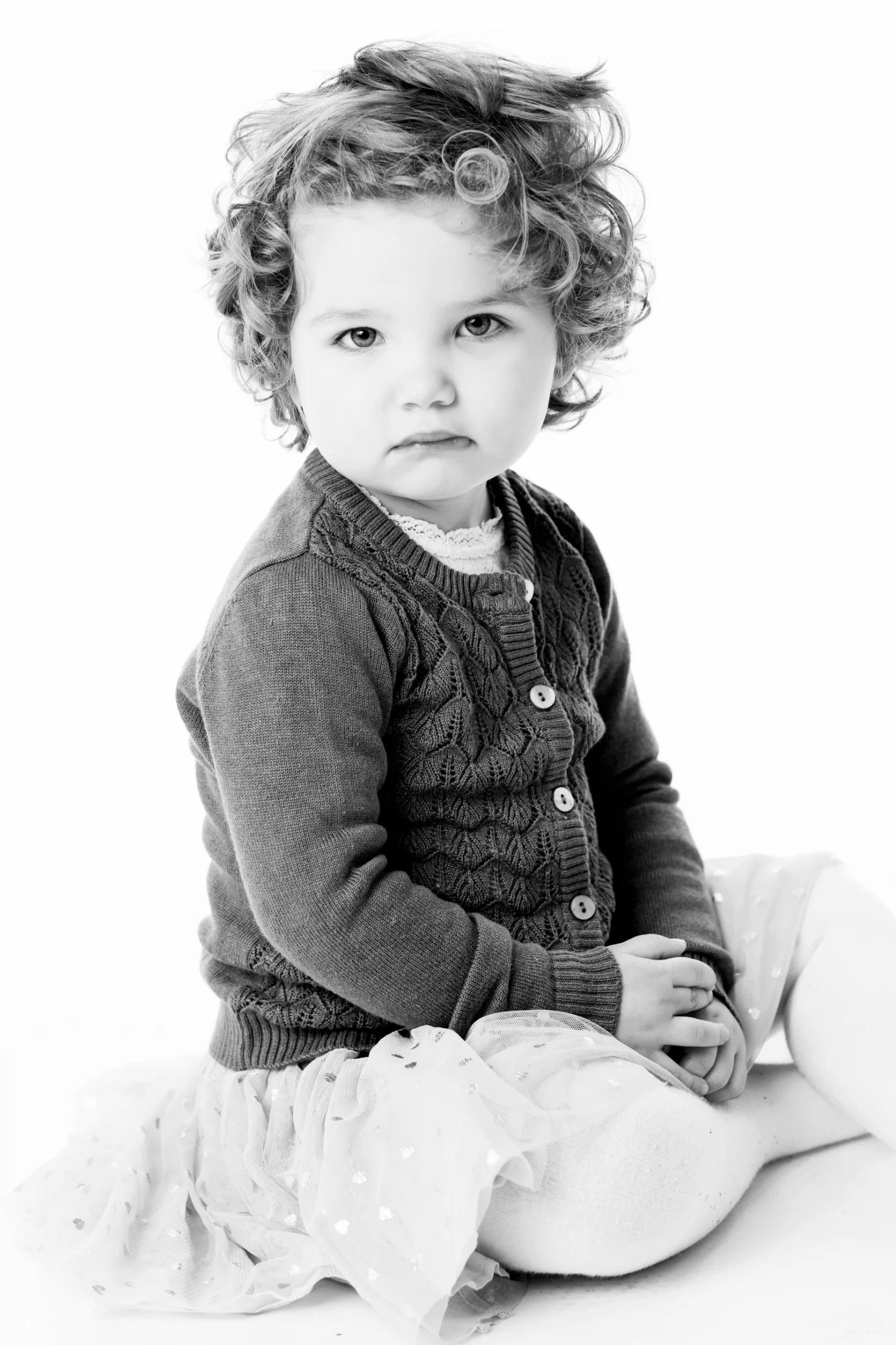 Sort-hvid fotografering af et lille barn med krøllet hår der sidder og kikker alvorligt på en hvid baggrund.
