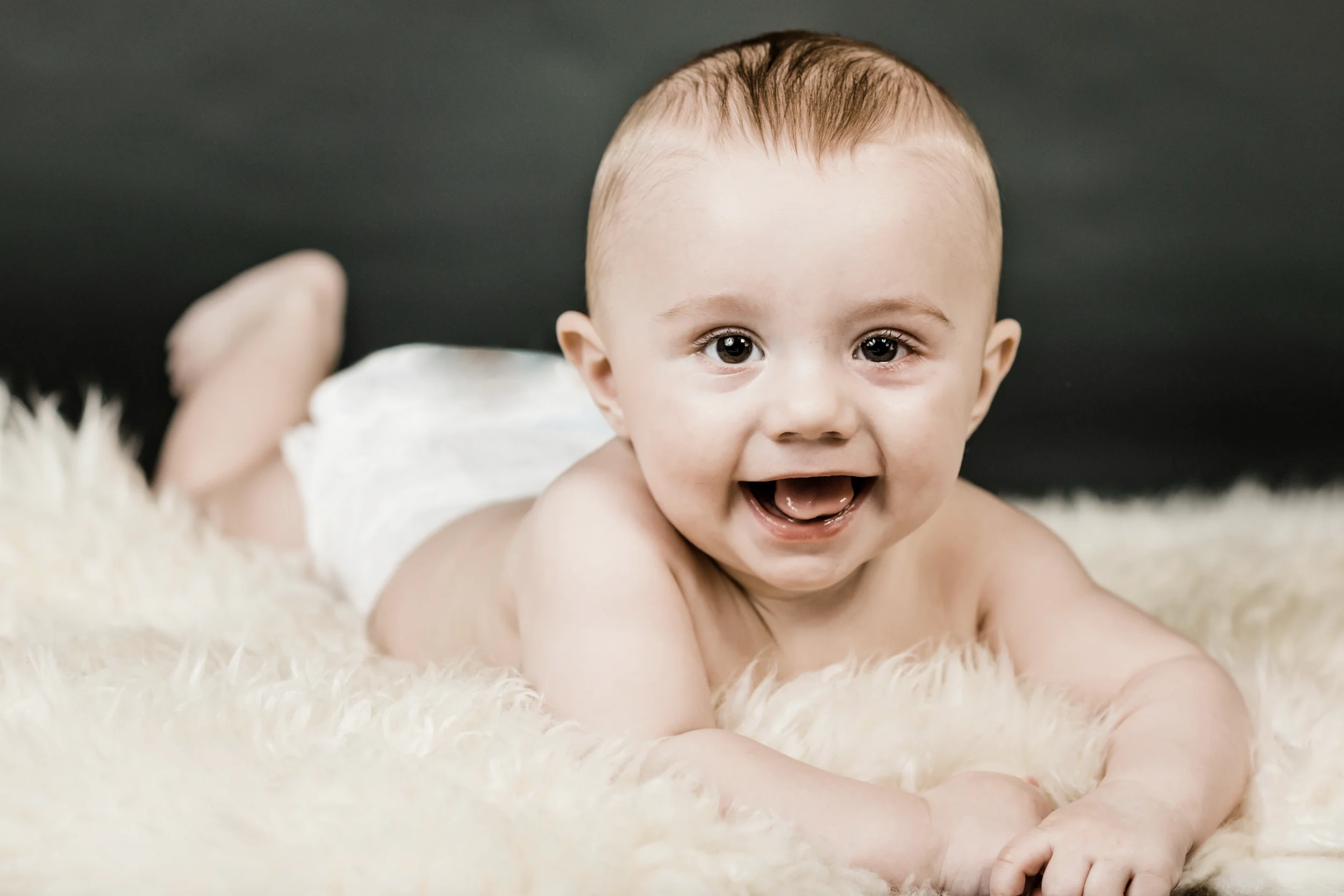 Glad baby ligger på maven på et hvidt pelstæppe med mørk baggrund, giver et stort smil til kameraet.