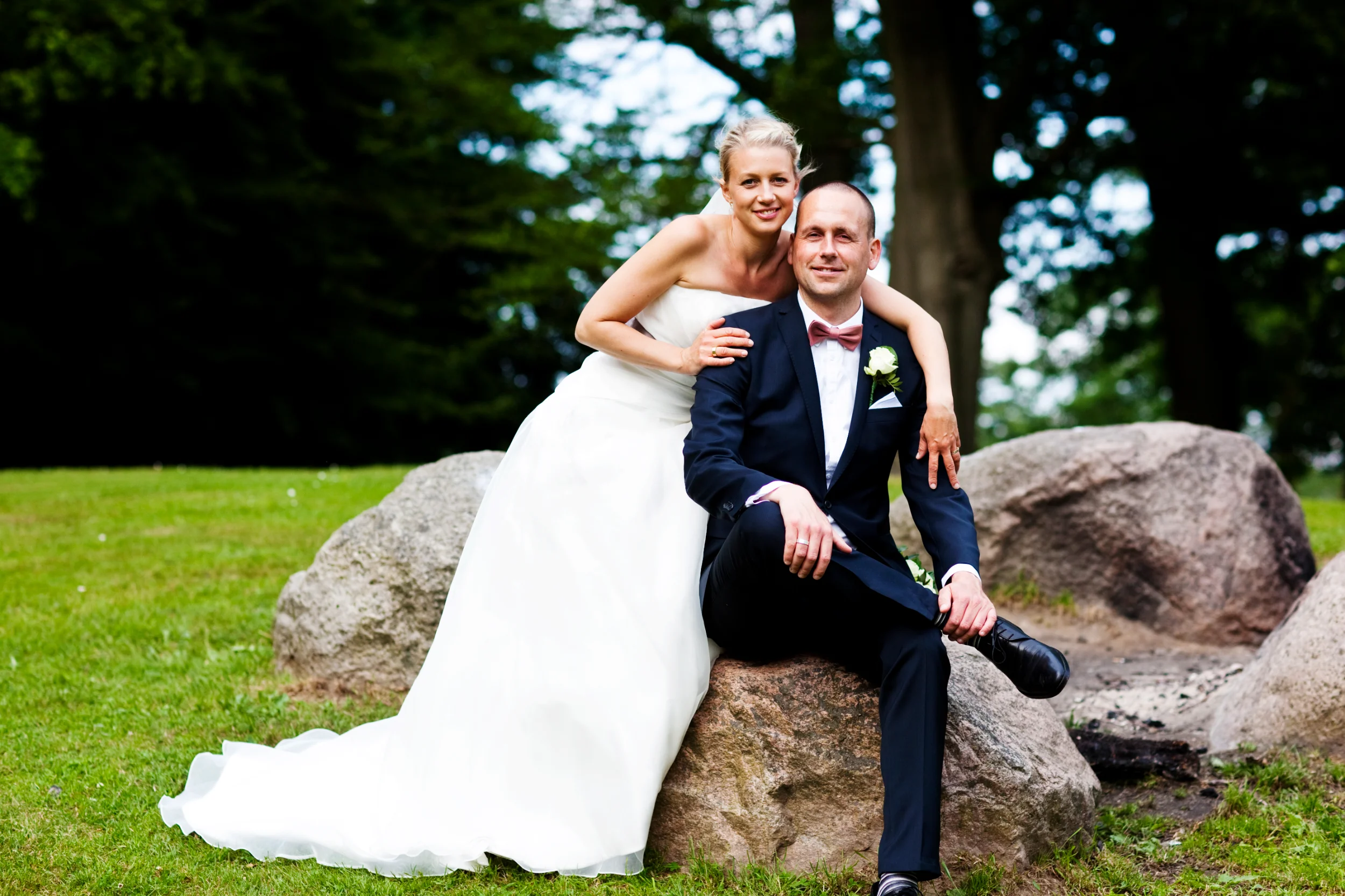 Bryllupsfoto af brudepar, der smiler til kameraet, hvor manden sidder på en sten og kvinden står bag ham i Virum.
