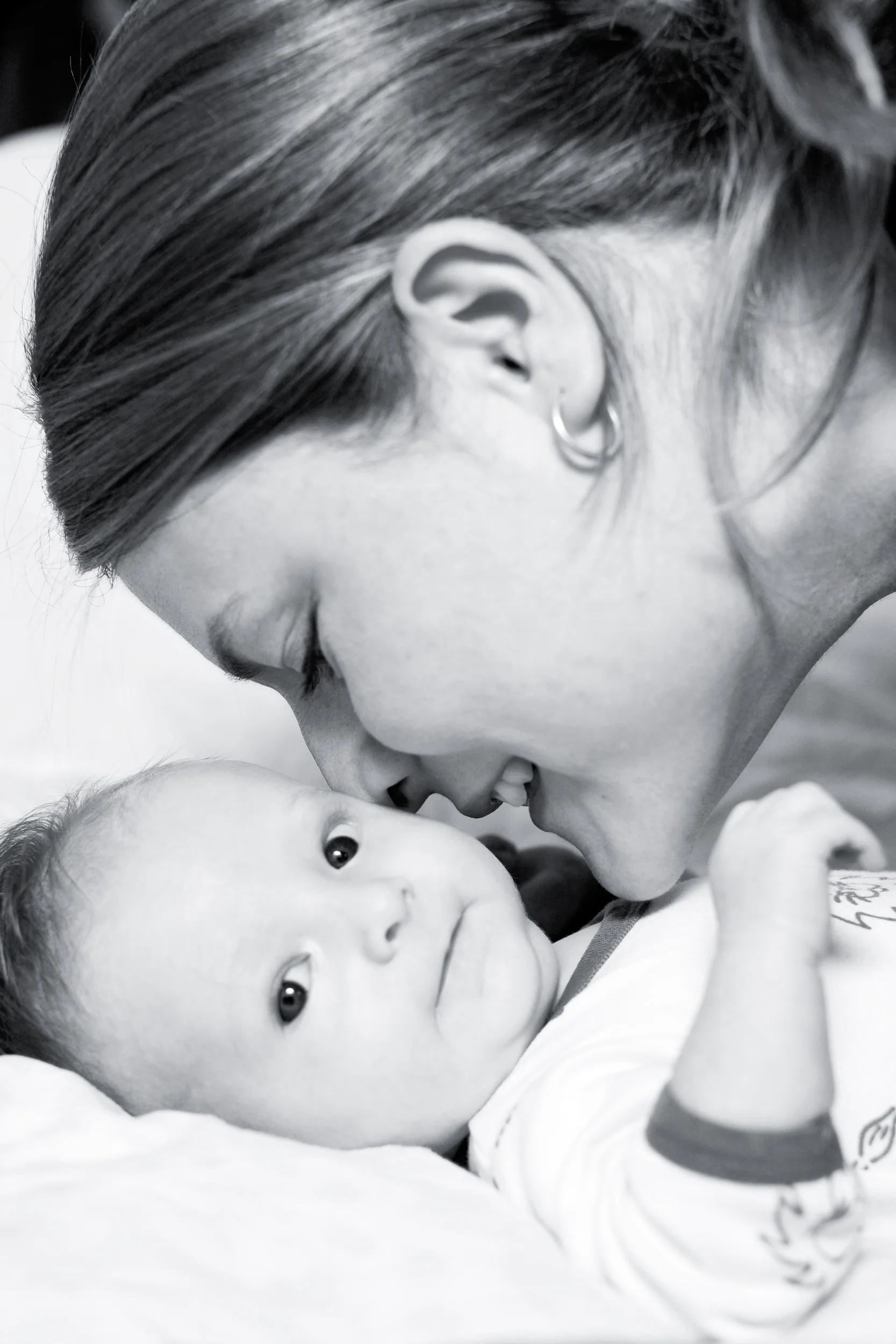 Sort/hvid babyfoto af mor der kysser sit barn på panden med rørende nærhed. Babyen kigger nysgerrigt frem.