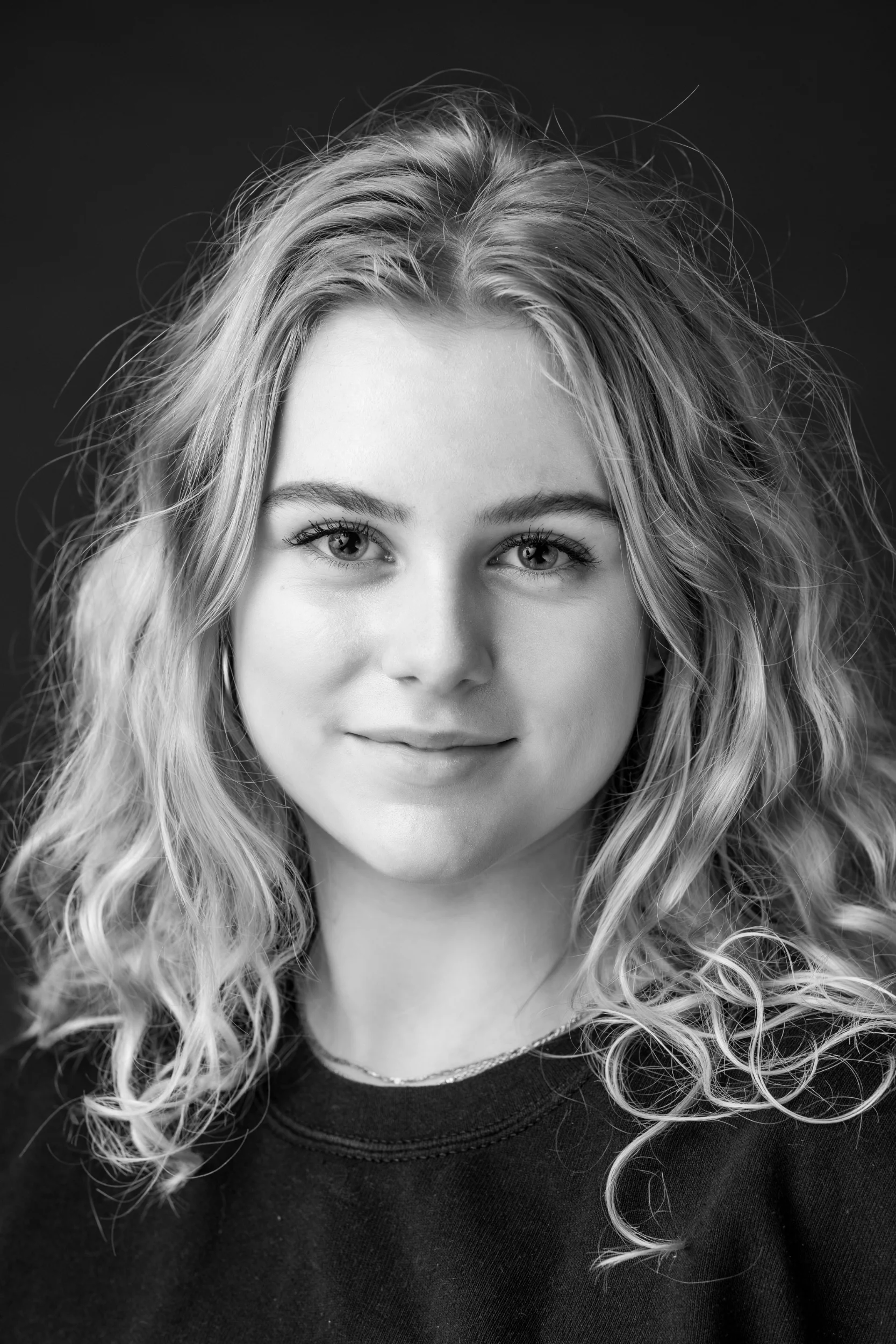 Sort/hvid portrætfoto af smilende, blond teenager med krøllet hår, der ser direkte ind i kameraet.