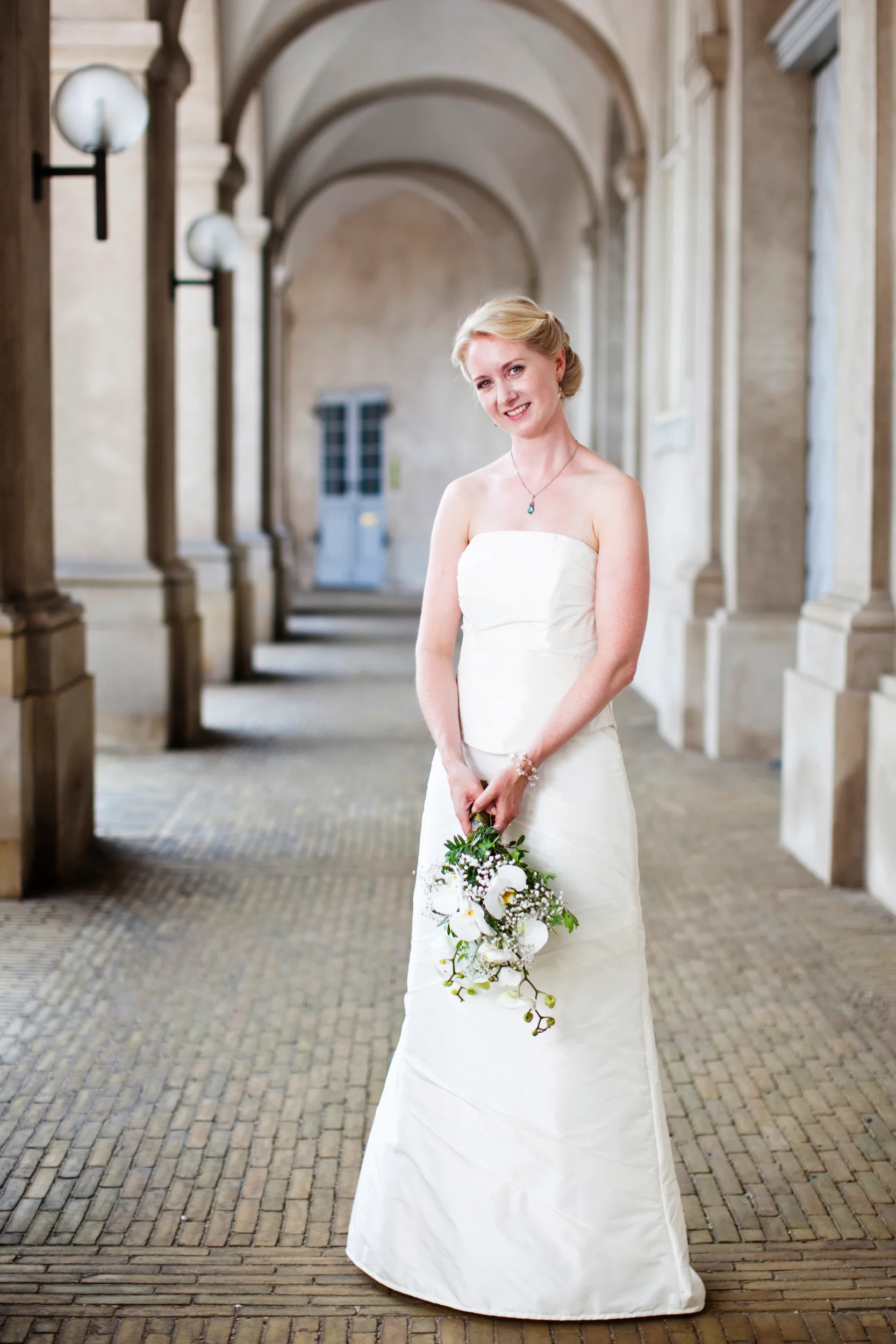 Bryllupsfotografering af smilende brud i hvid kjole med brudebuket på Christiansborg Ridebane. Lyse, glade farver.