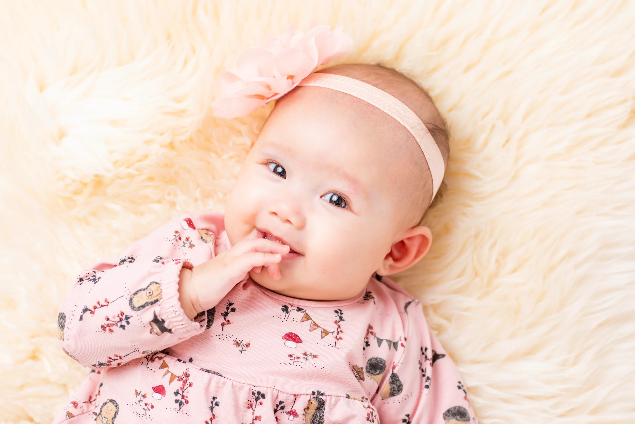 Nærbillede af smilende baby i en lyserød kjole med et blomstret hårbånd, liggende på et lunt, lyst tæppe. Babyfotografering.