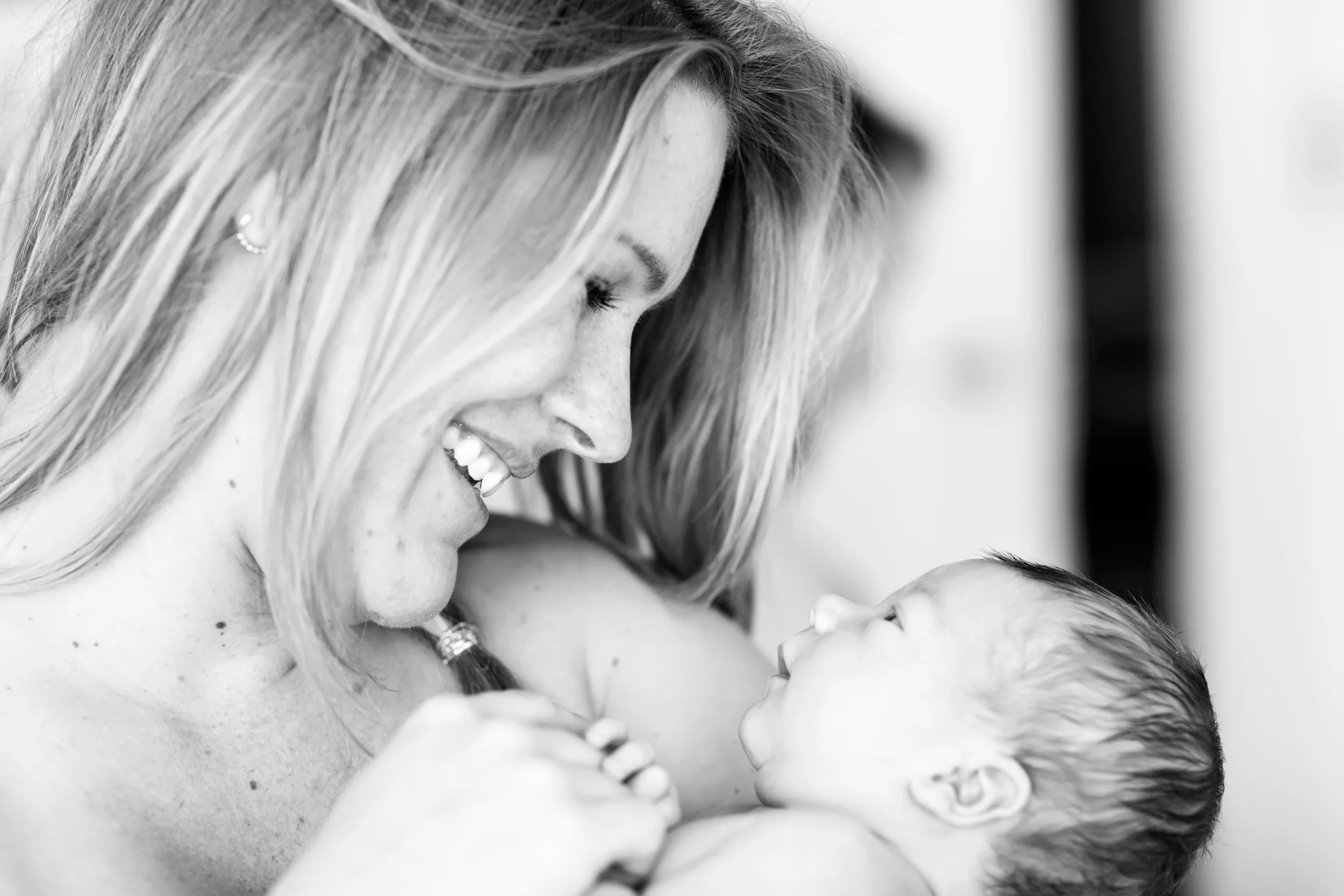 Babyfotografering i sort/hvid af smilende mor, der kigger kærligt på sin nyfødte baby. Blødt lys skaber intim stemning.