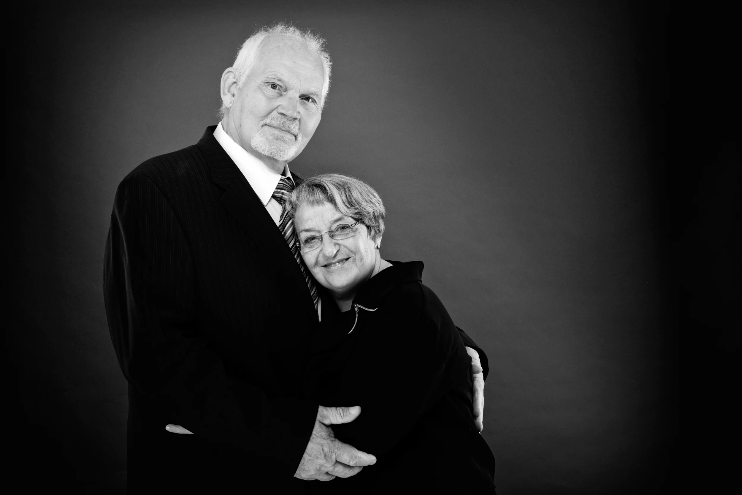 Sort-hvidt portrætfoto af smilende ægtepar. Han i jakkesæt omfavner hende, der læner sig ind mod ham og smiler til fotogra...
