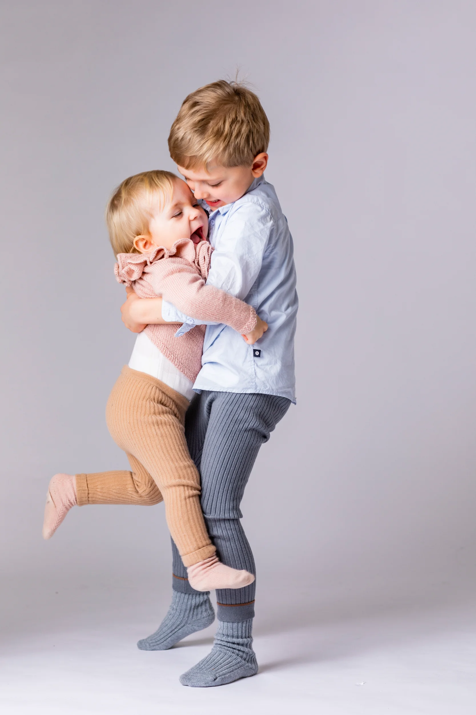 Børn: En glad dreng giver sin lillesøster et varmt kram. Hun griner med åben mund, mens han holder og løfter hende blidt.