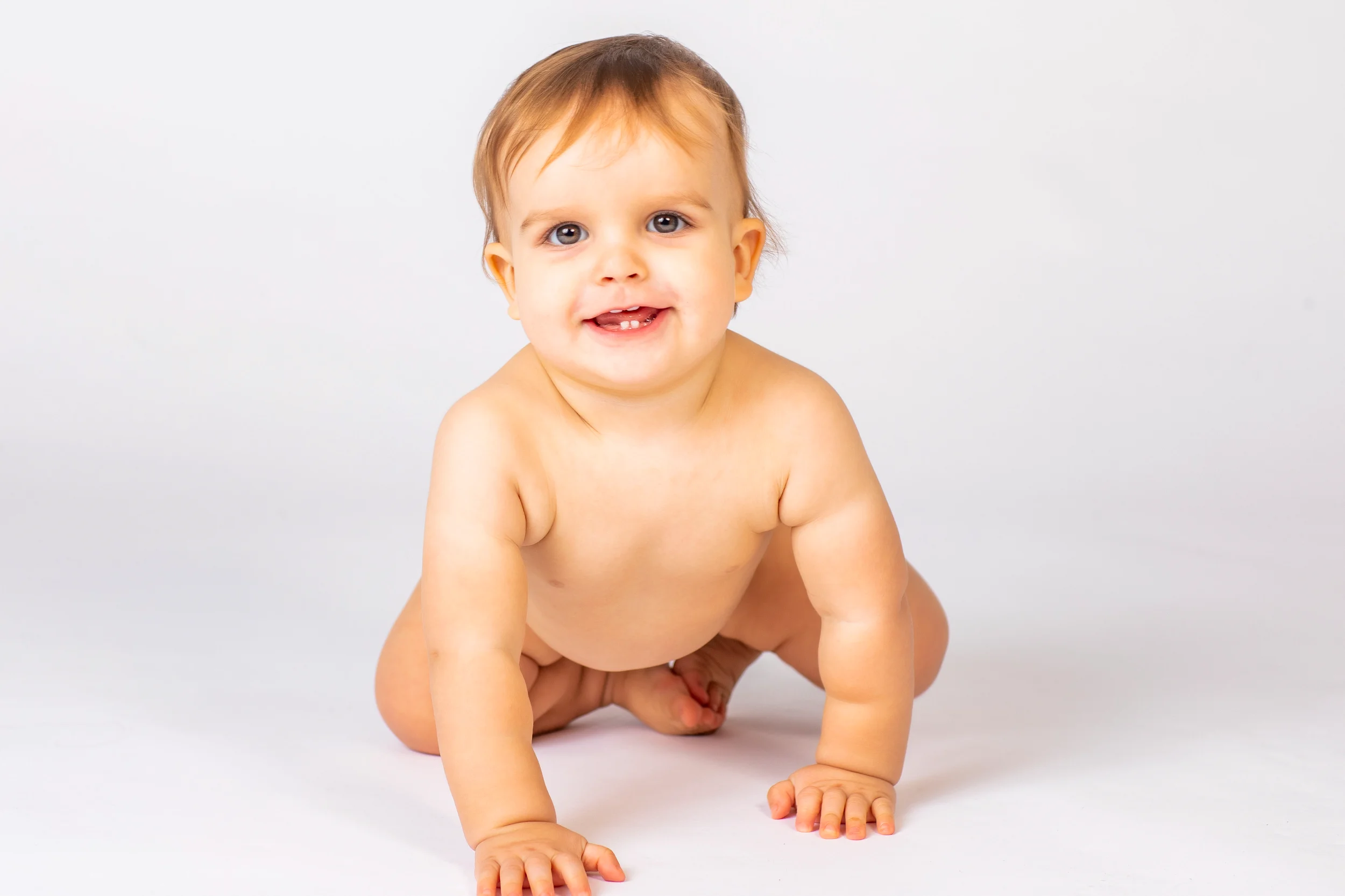 Glad baby kravler og smiler mod beskueren. Autentisk babyfotografering med hvid baggrund i naturligt lys.