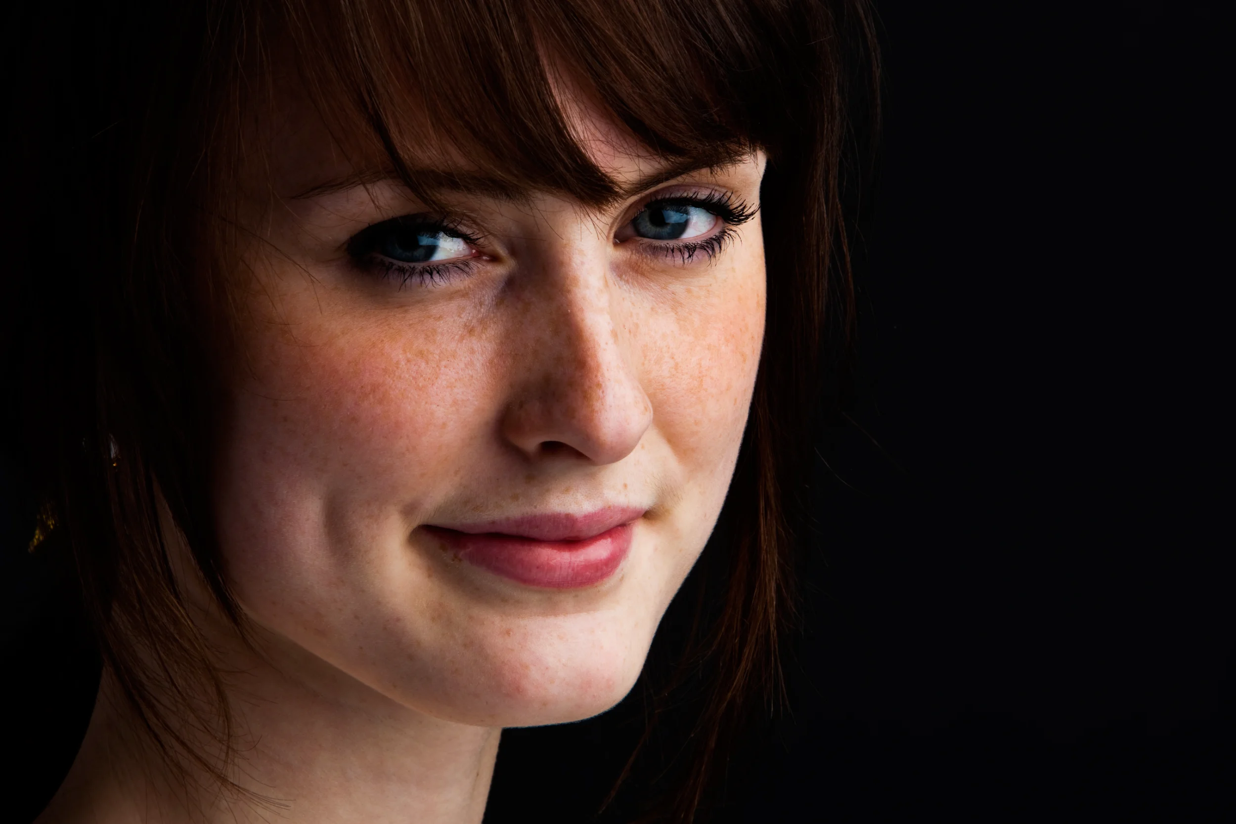 Nærbillede af ung kvinde med fregner, brunt hår og et varmt smil. Vinklen understreger personligheden i dette portrætfoto.