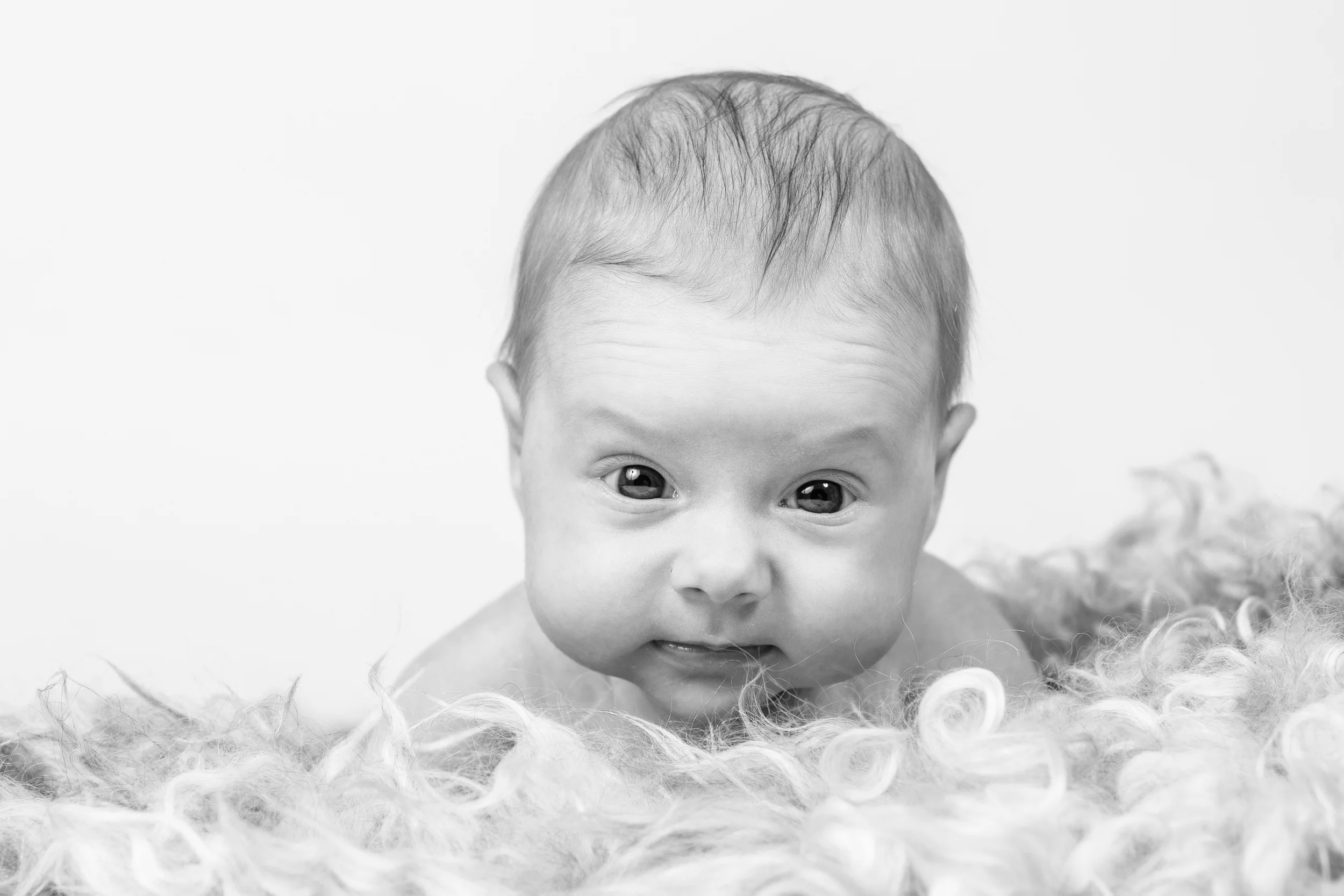 Babyfotografering: Sort-hvidt nærbillede af nysgerrig baby der kigger frem fra et blødt underlag.