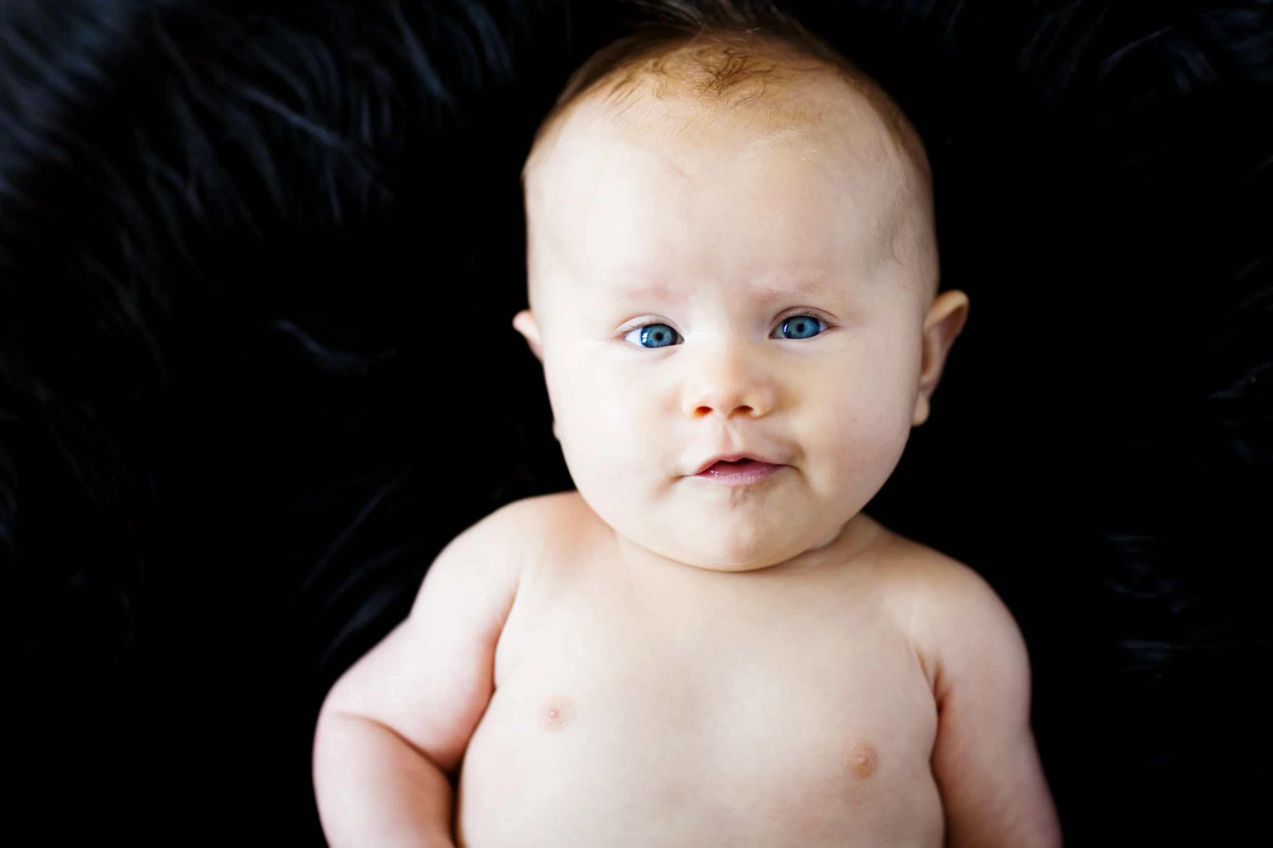 Nærbillede af baby med klare blå øjne liggende på en blød, mørk baggrund til et personligt babyfotografi.