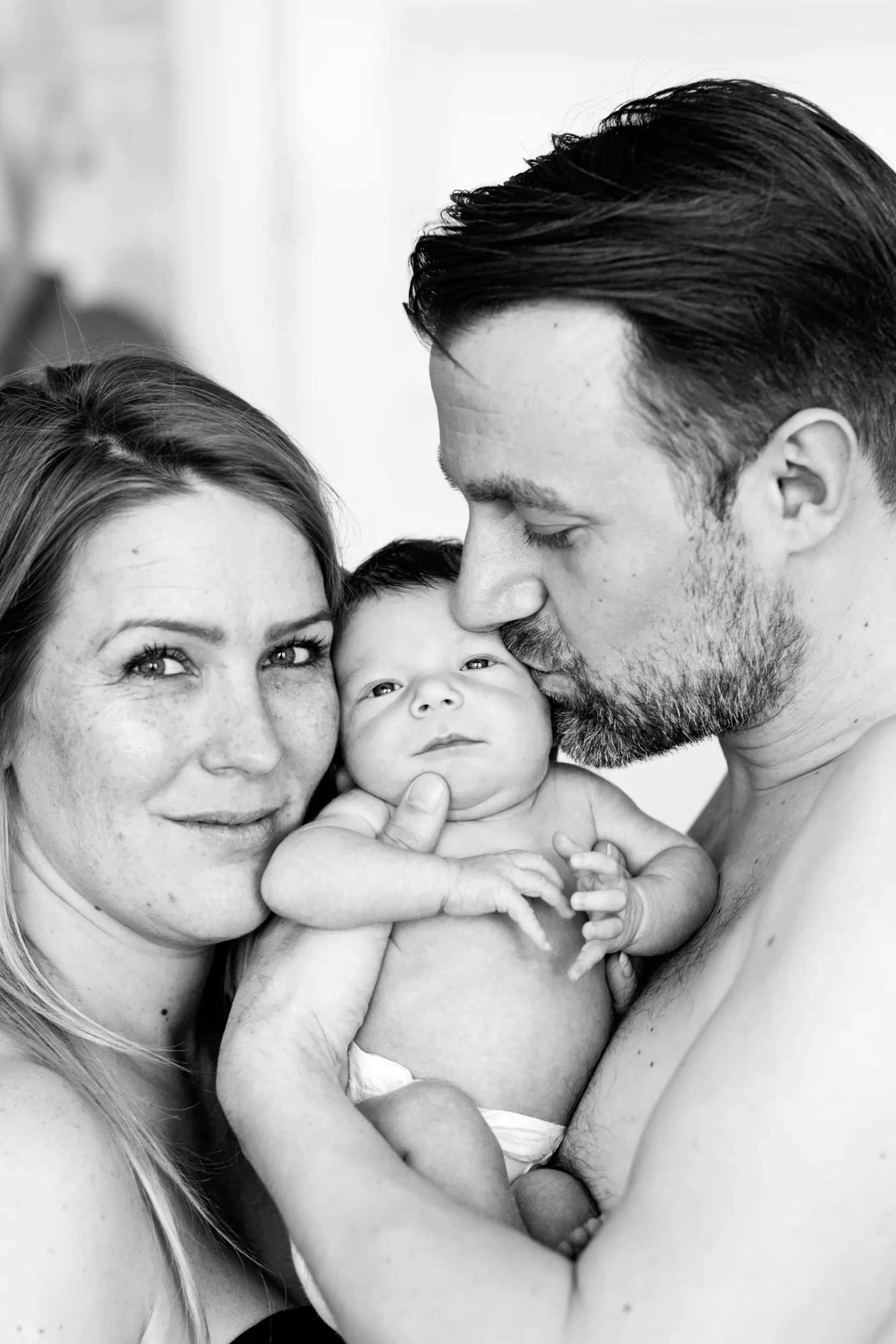 Sort-hvid babyfotografering af forældre, der holder deres nyfødte. Faren kysser babyens pande, moren smiler til kameraet.