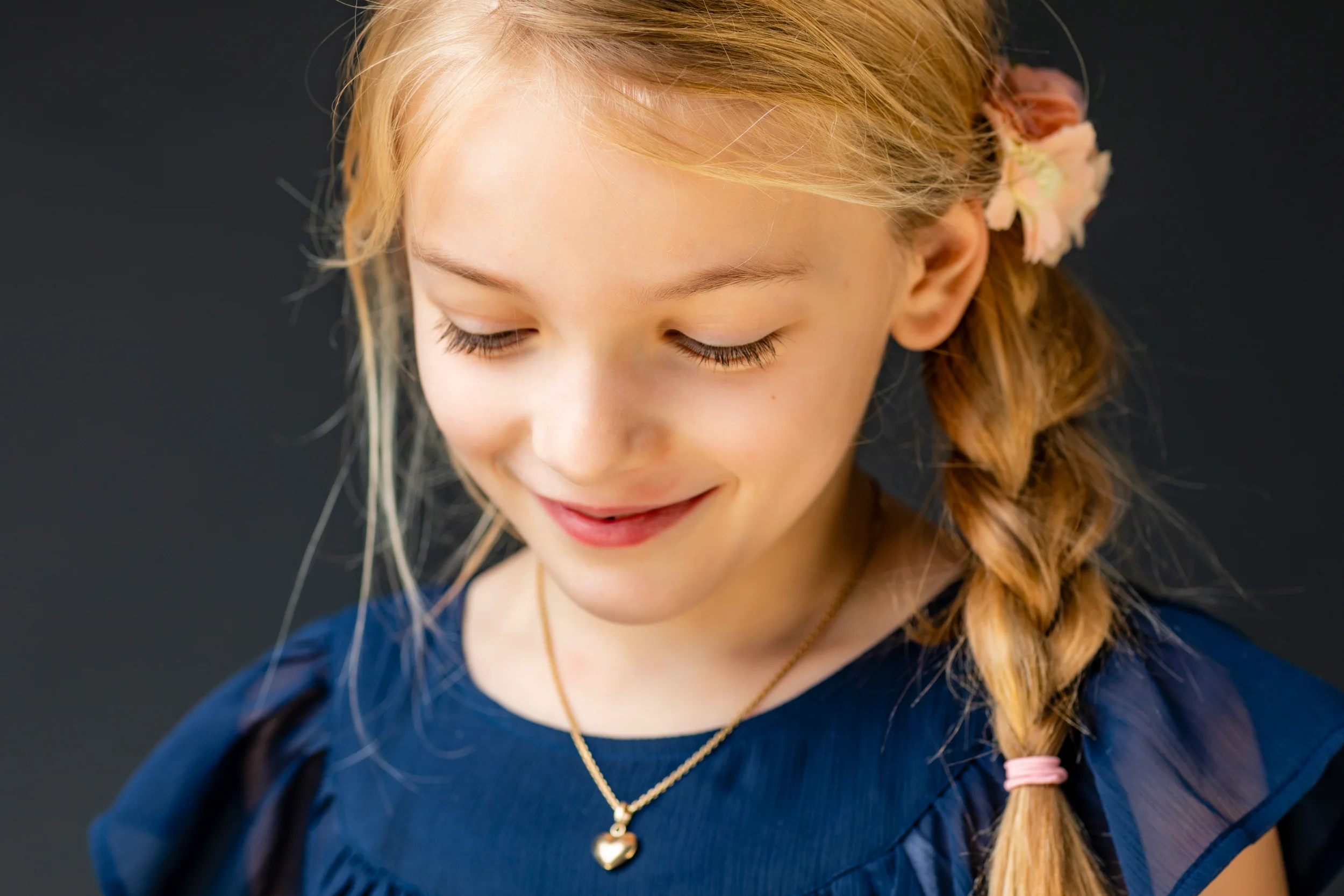 Børnefotografering af en pige med fletning, nedslåede øjne med smil på læben, iført en blå overdel og guldhalskæde.