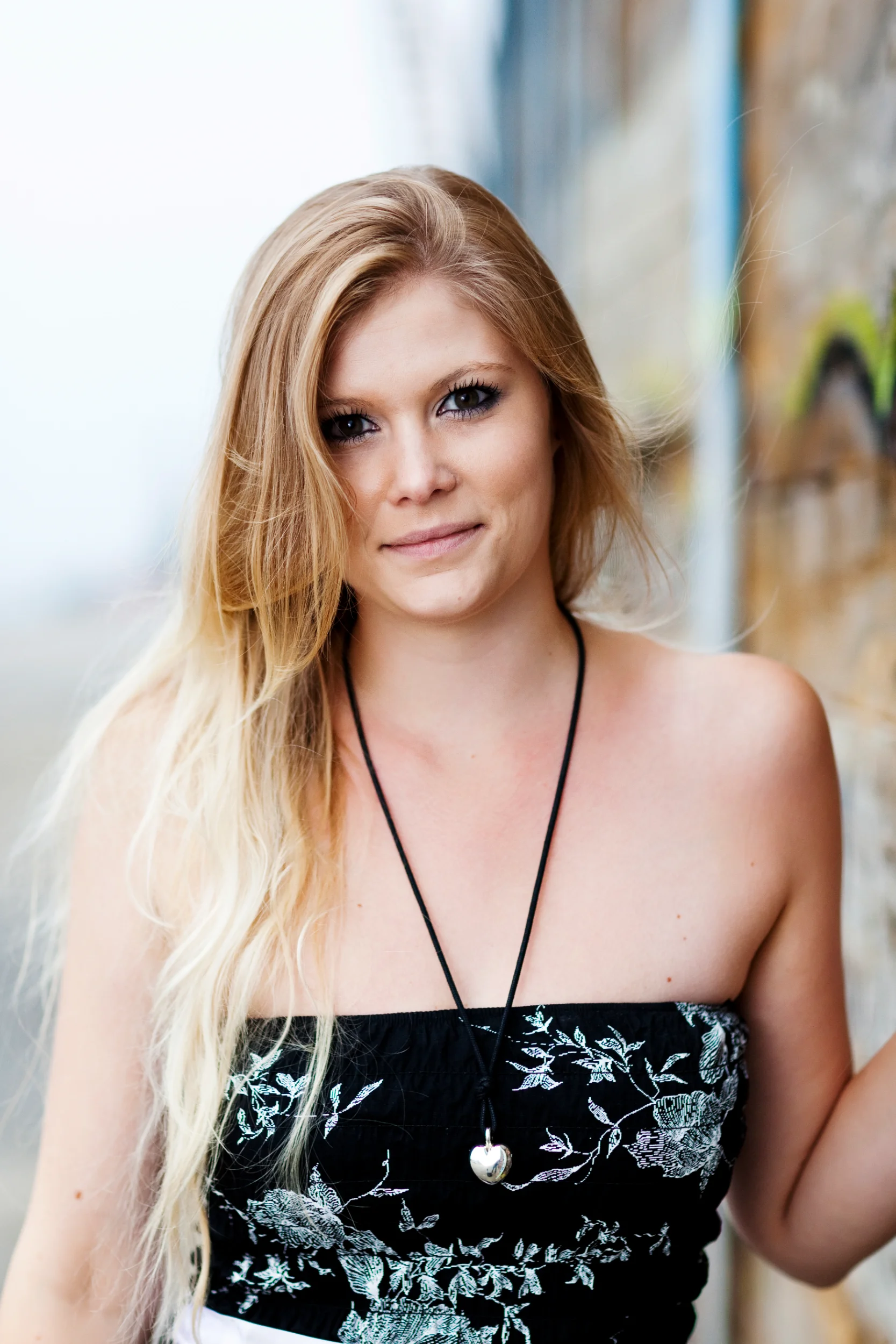 Portrætfoto af smilende ung kvinde med langt, lyst hår og sort stropløs top, der står op ad en væg i dagslys.