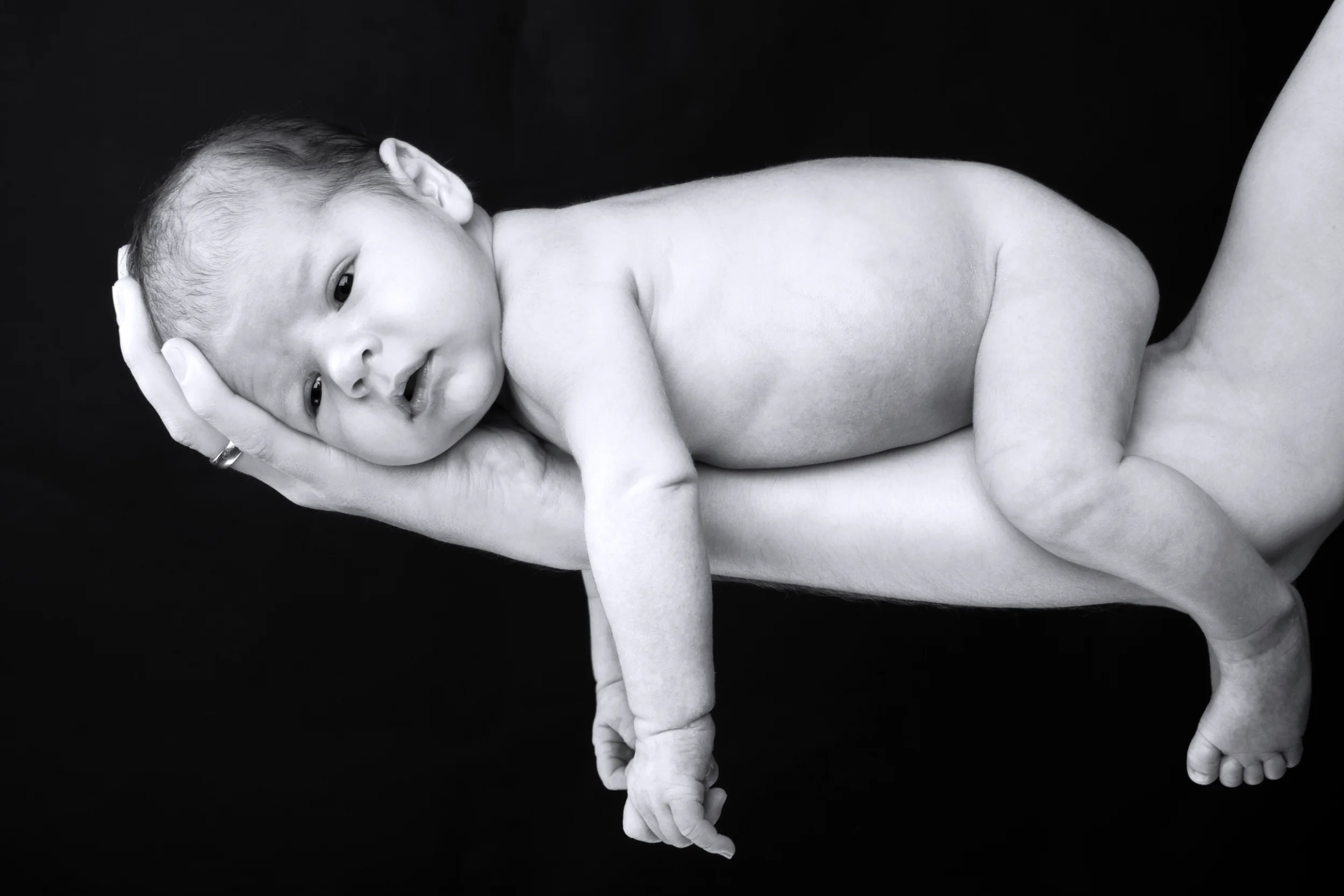 Nyfødt fotografering hvor en baby hviler trygt og nøgen i voksne hænder. S/H, fredfyldt stemning med blødt lys.
