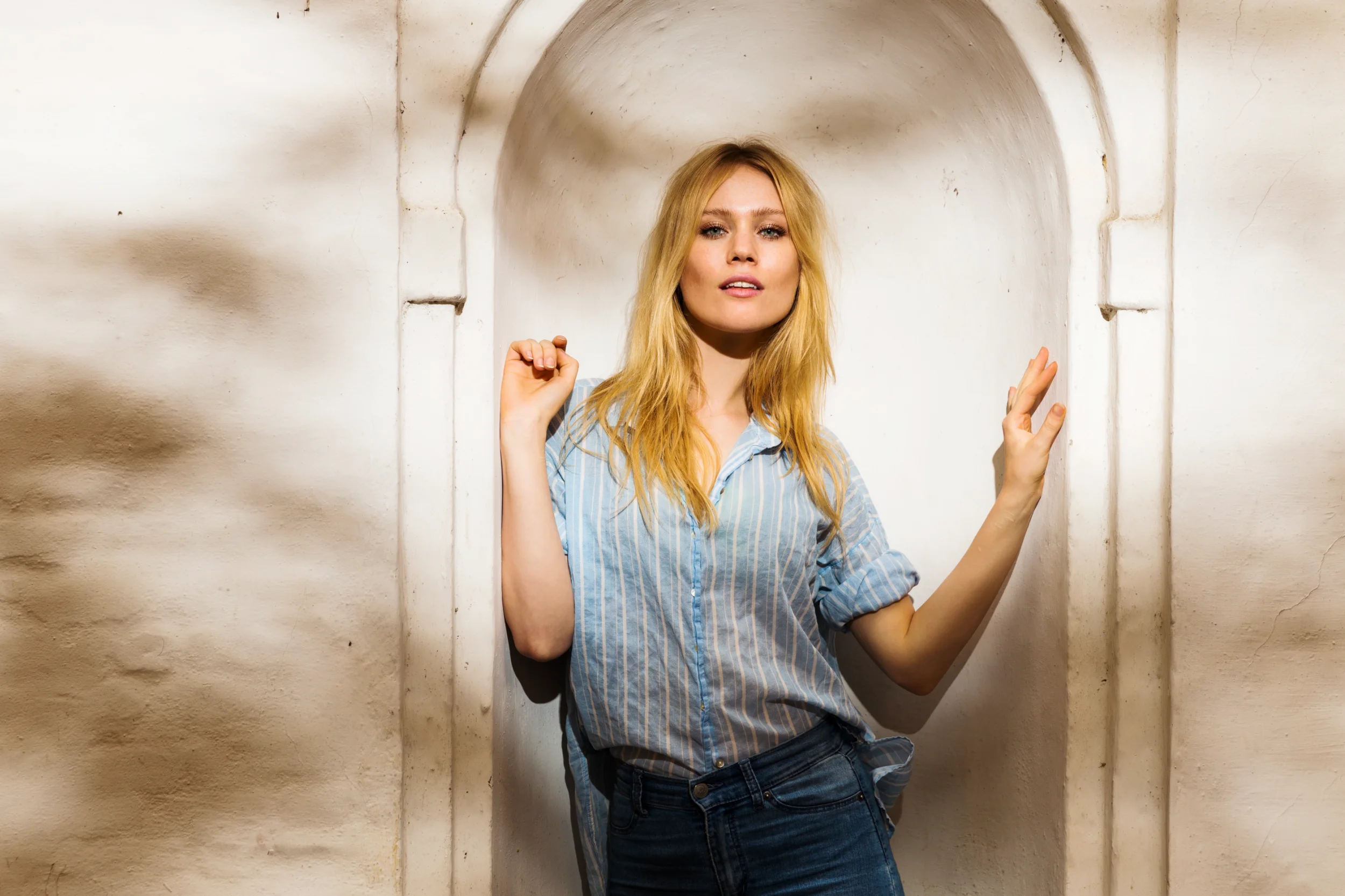 Portrætfoto af smuk kvinde med blondt hår i stribet skjorte, der står i en buet niche med spændende skygger.