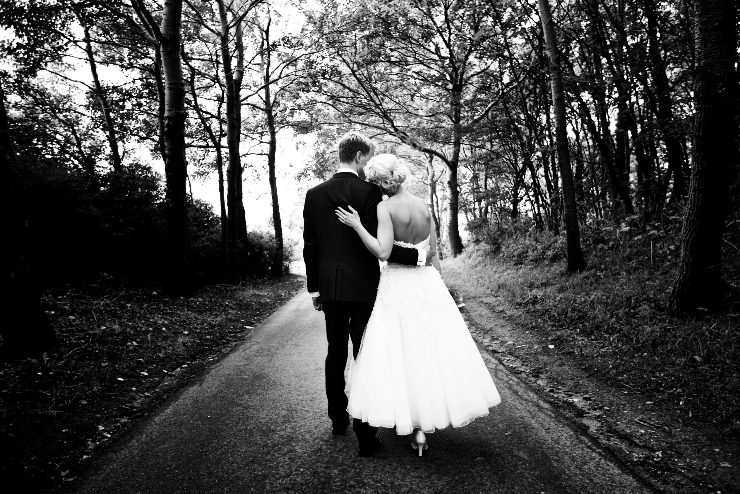 Bryllupsfotografering: Sort/hvidt foto af brudepar, set bagfra, der går arm i arm på en vej i en skov med højtsluttende tr...