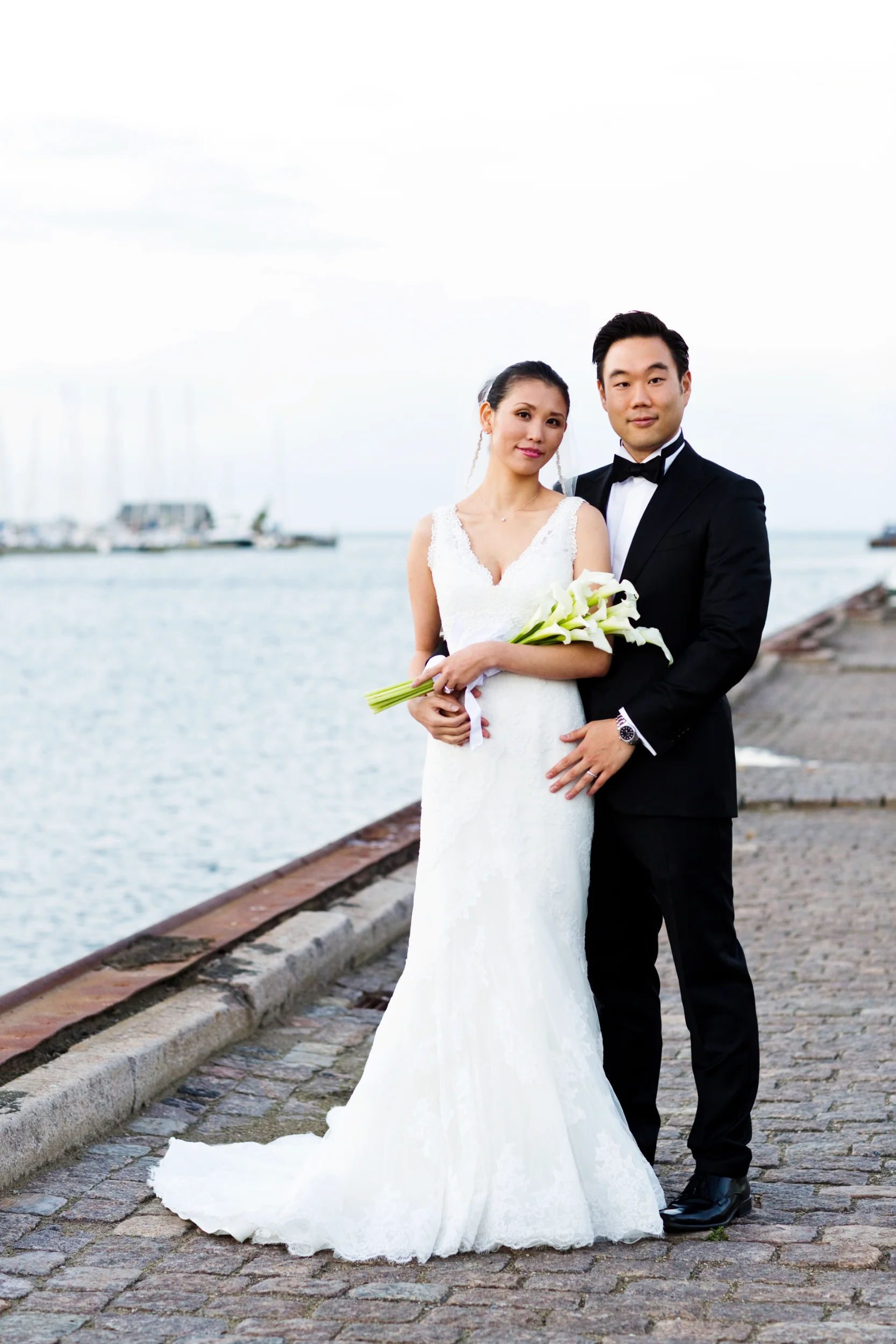 Bryllupsfoto af smilende par ved vandet i Nordhavn. Brud i hvid kjole, gom i sort smoking, holder buket med hvide kallaer.
