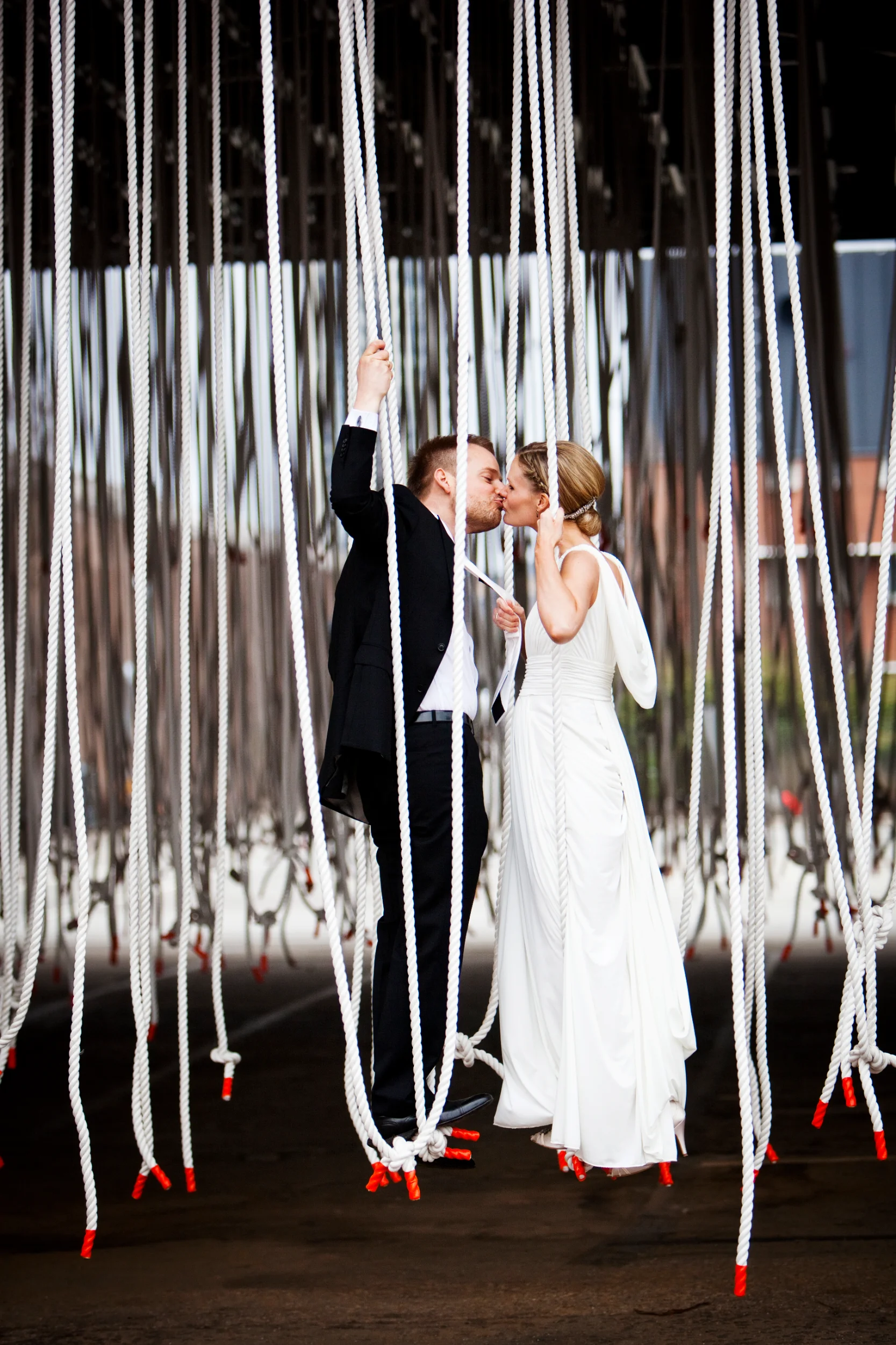 Bryllupsfotografi: Brudepar kysser i et rebstårn med mange lodrette reb. Sort/hvidt billede med fokus på glæde.