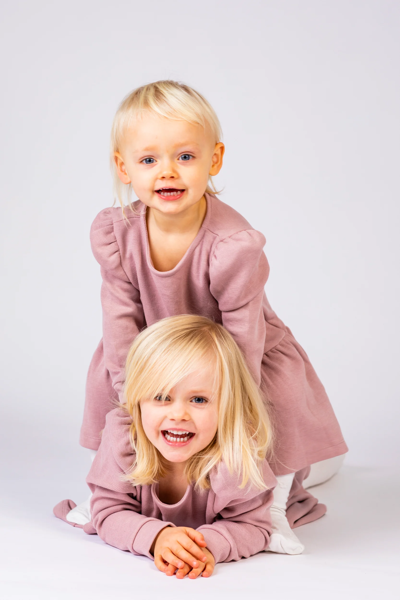 To legende blonde børn i rosa tøj smiler stort mod kameraet under en fotografering med hvid baggrund.