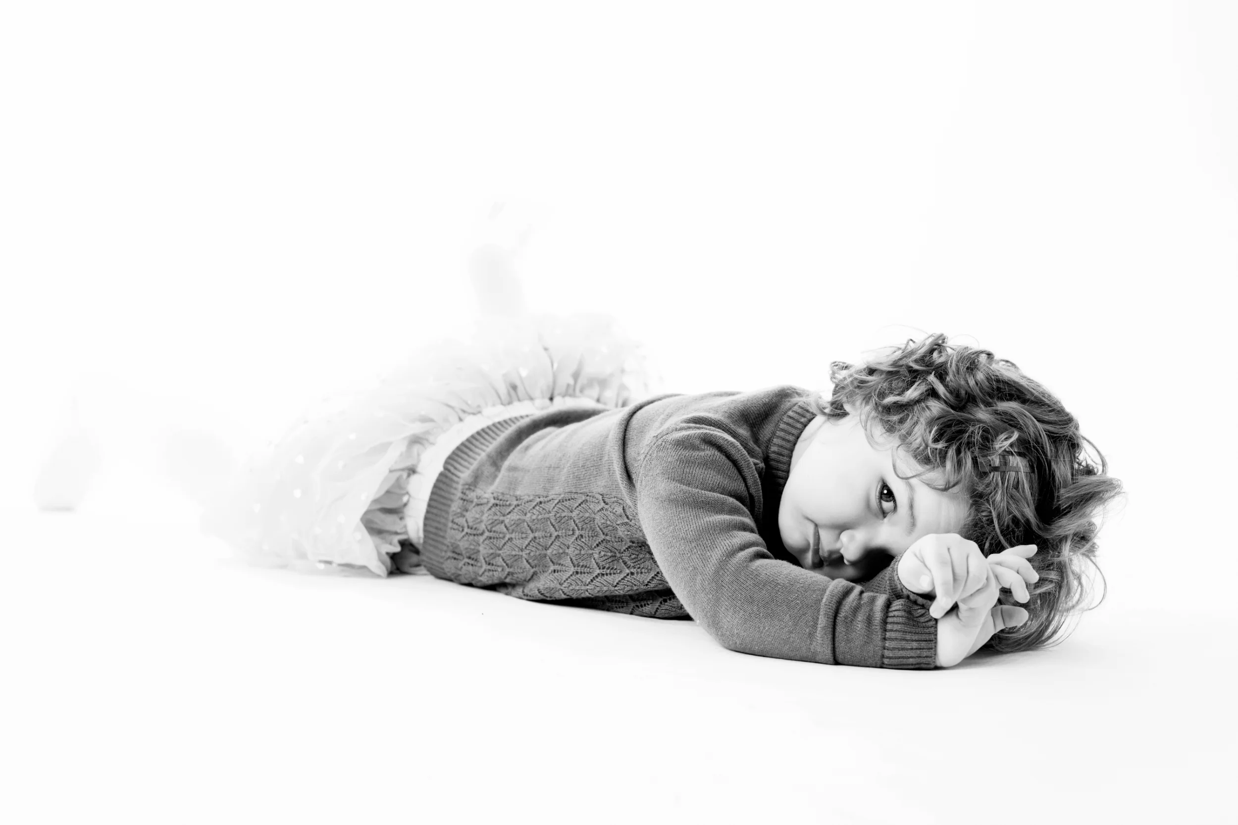 Sort-hvidt børnefoto af en lille pige med krøllet hår, der hviler på maven og kigger direkte ind i kameraet.