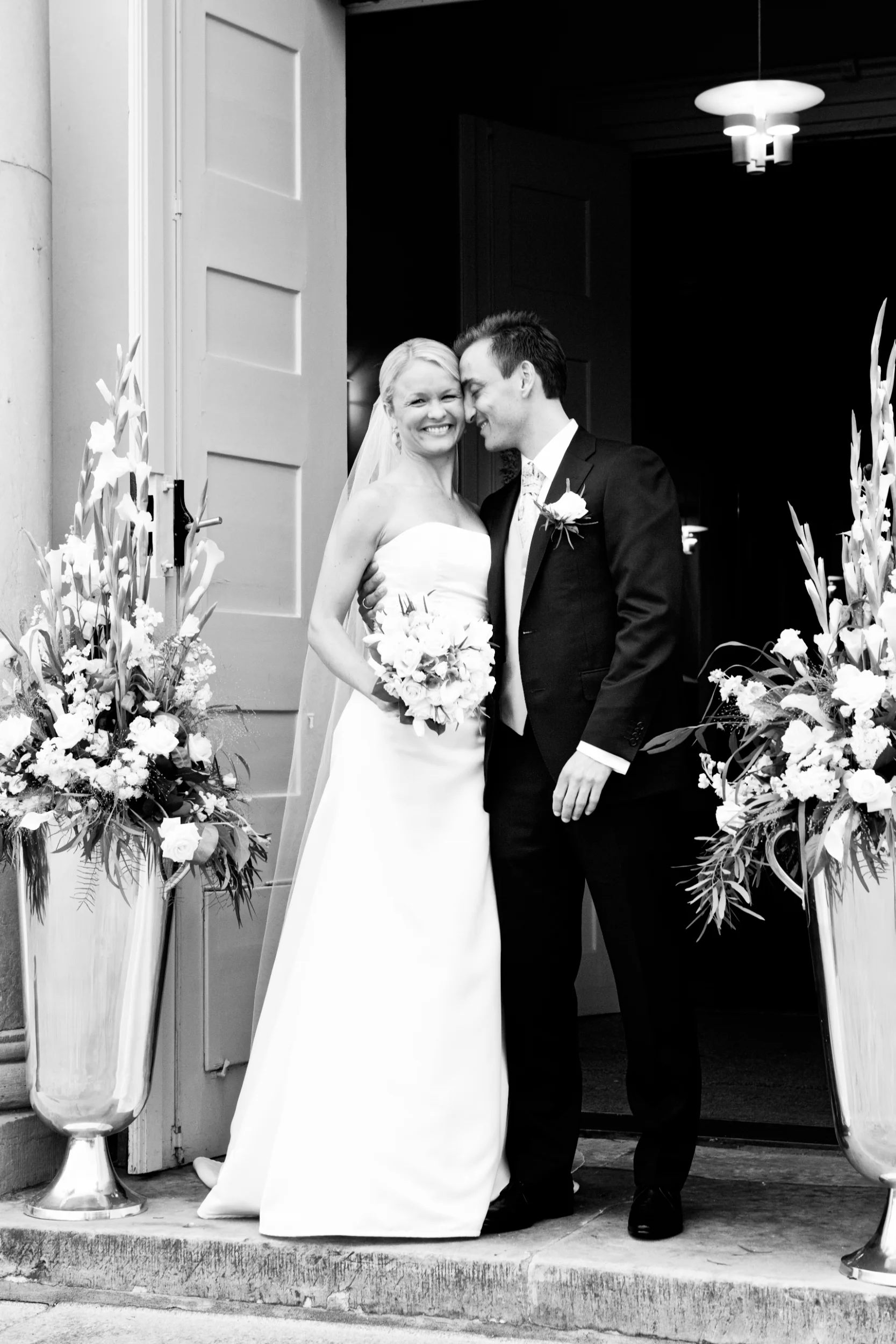 Bryllupsfotografering: Smilende brudepar i omfavnelse foran kirken i Virum, omgivet af hvide blomsterarrangementer.