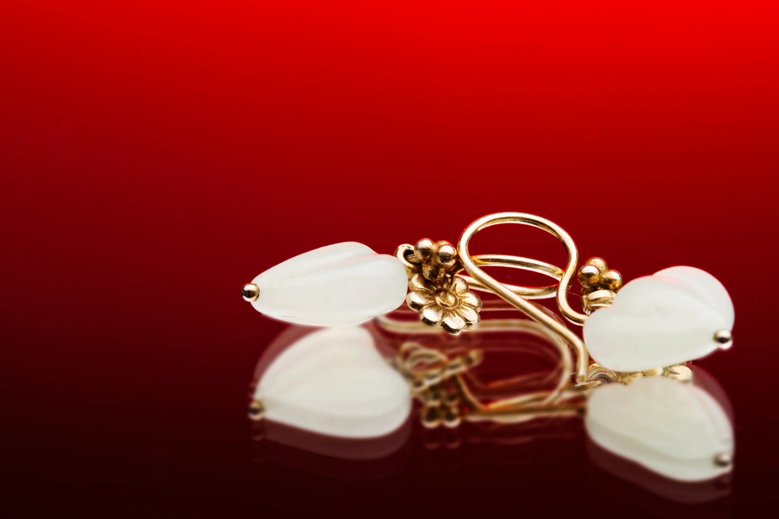 Nærbillede af et par elegante guldøreringe med hvide perler, der reflekteres på en blank rød baggrund. Andet fotografering.