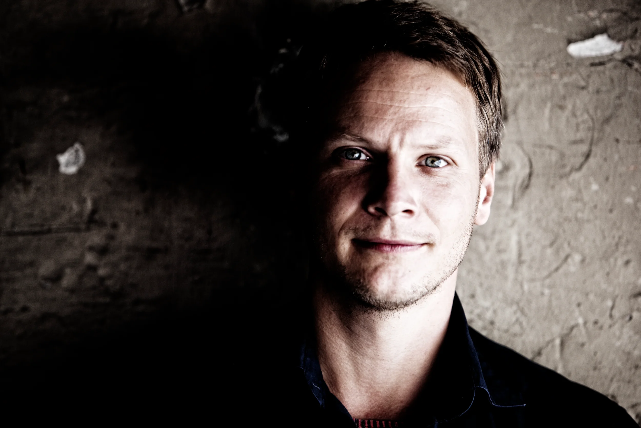 Portrætfoto af ung mand med blåt øje og mørkt hår, delvist belyst fra venstre mod en grov væg i dæmpet belysning.
