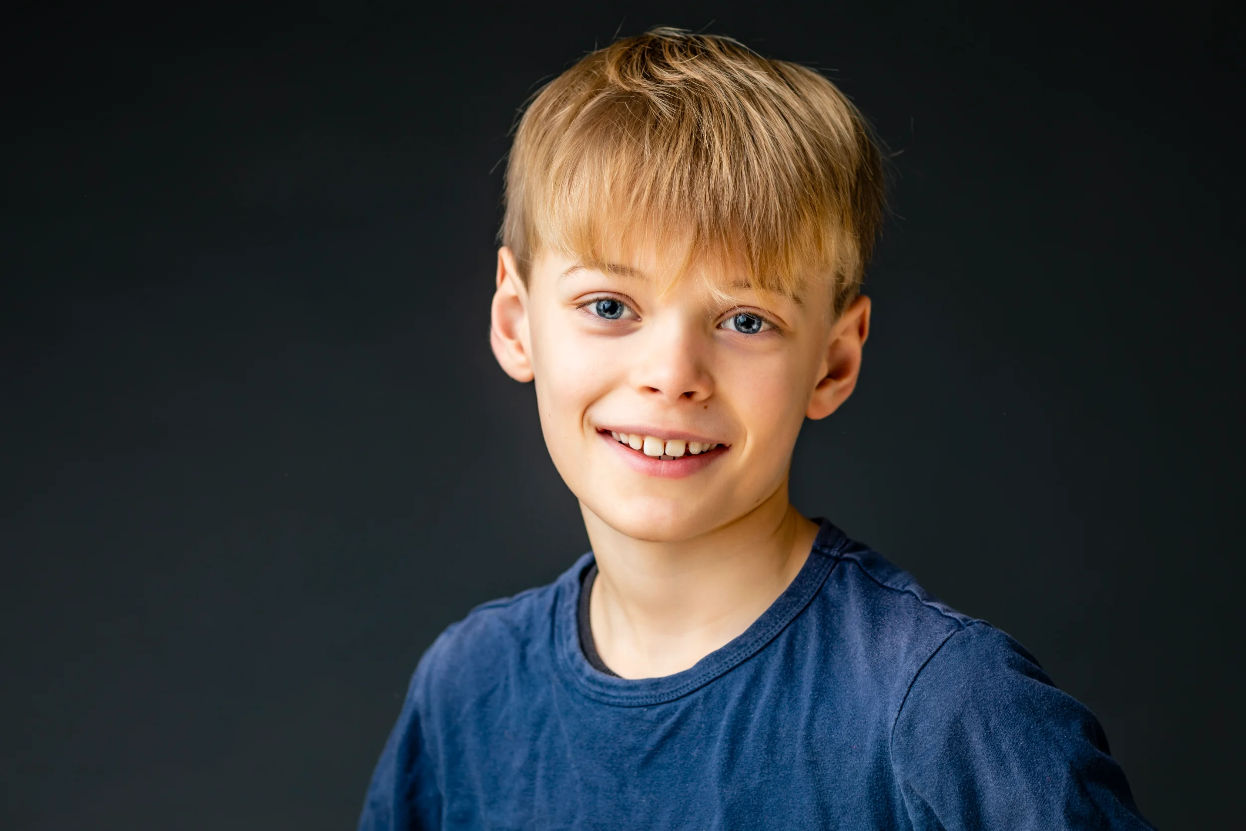 Børnefotografering: Smilende dreng med lyst hår og blå øjne i blødt lys, iført blå t-shirt.