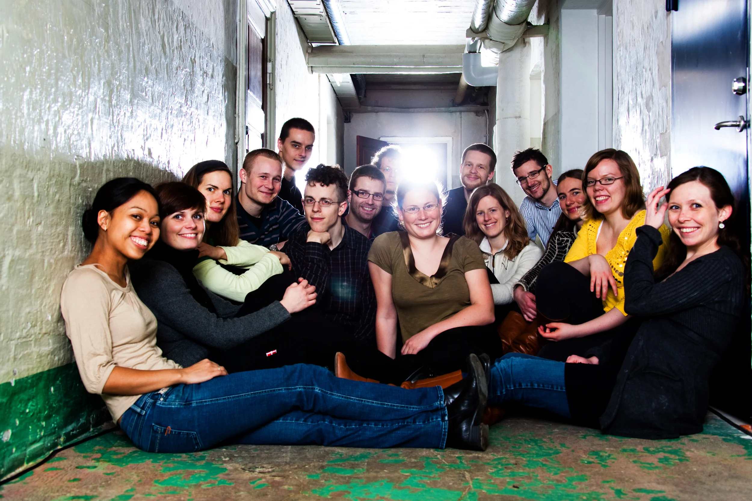 Gruppe af unge sidder smilende på gulvet i en smal gang med skarpt lys i baggrunden, et andet fotobillede.