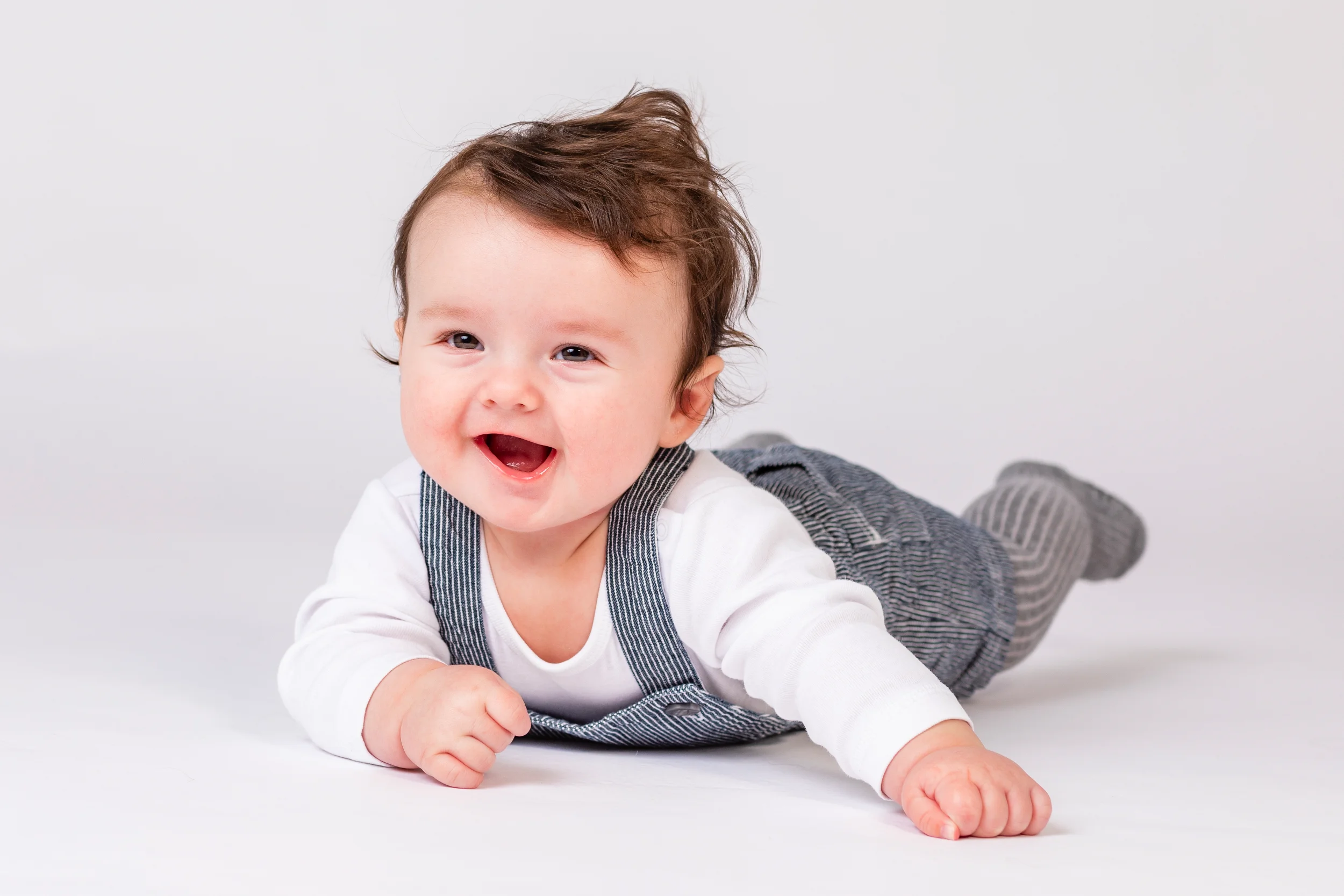 Glad baby i stribede seler og hvid trøje kravler mod beskuer i naturligt lys. Babyfotografering med fokus på glæde.
