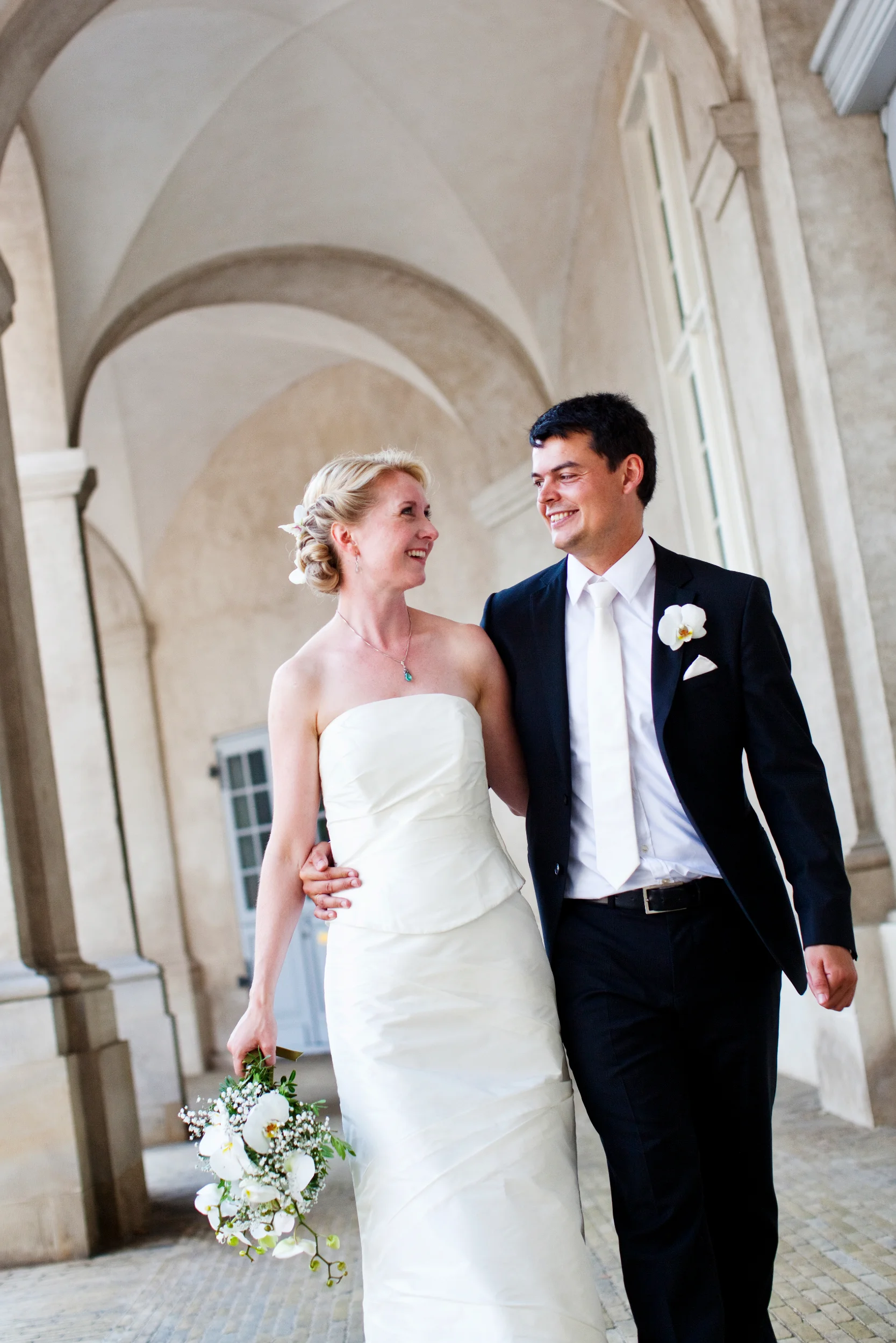 Bryllupsfotografi af glad brudepar, der går arm i arm under en buegang med et kærligt blik.