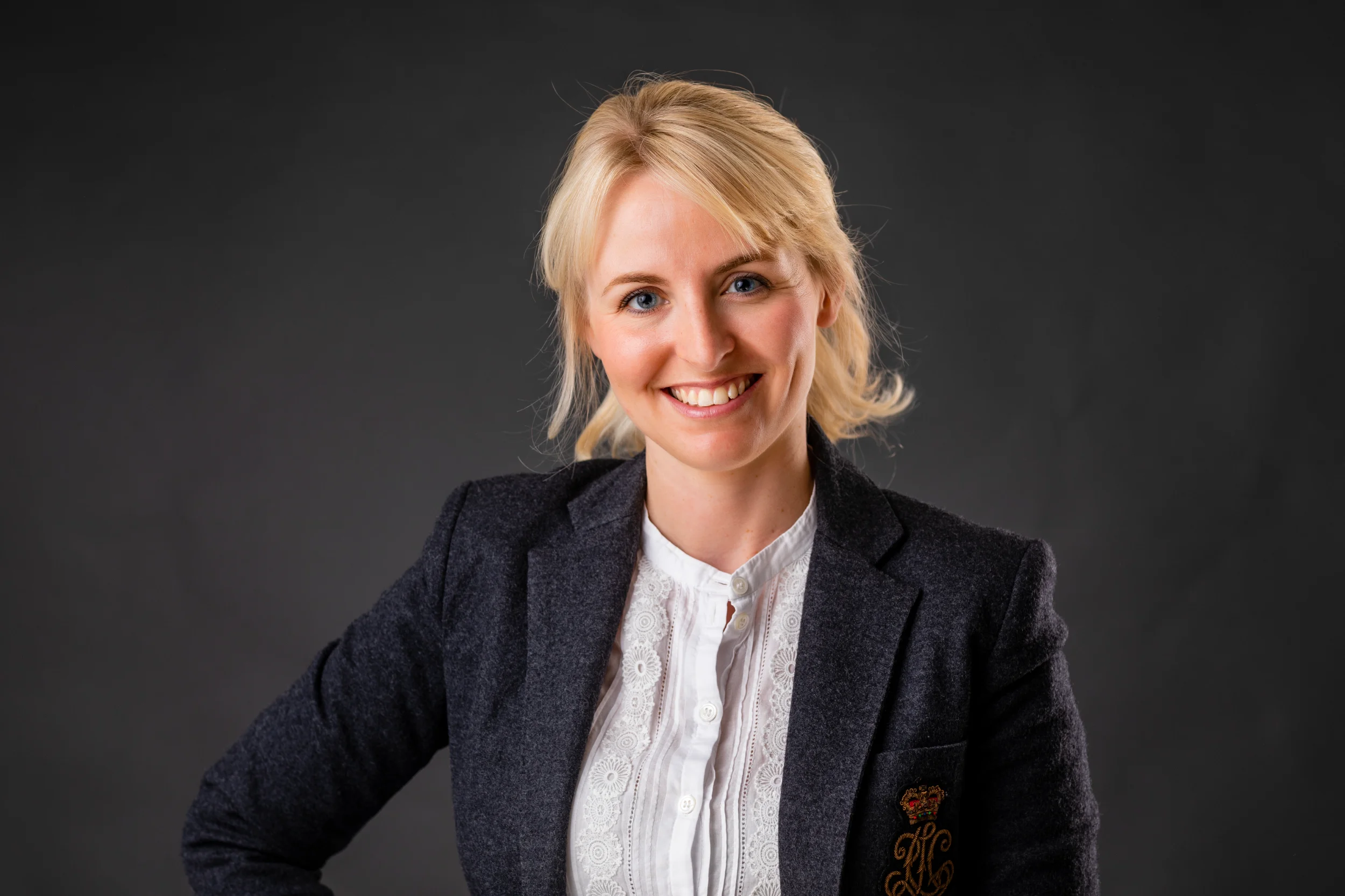 Portrætfoto af smilende kvinde med blondt hår i Virum iført mørk blazer og hvid blondeskjorte, mørk baggrund.