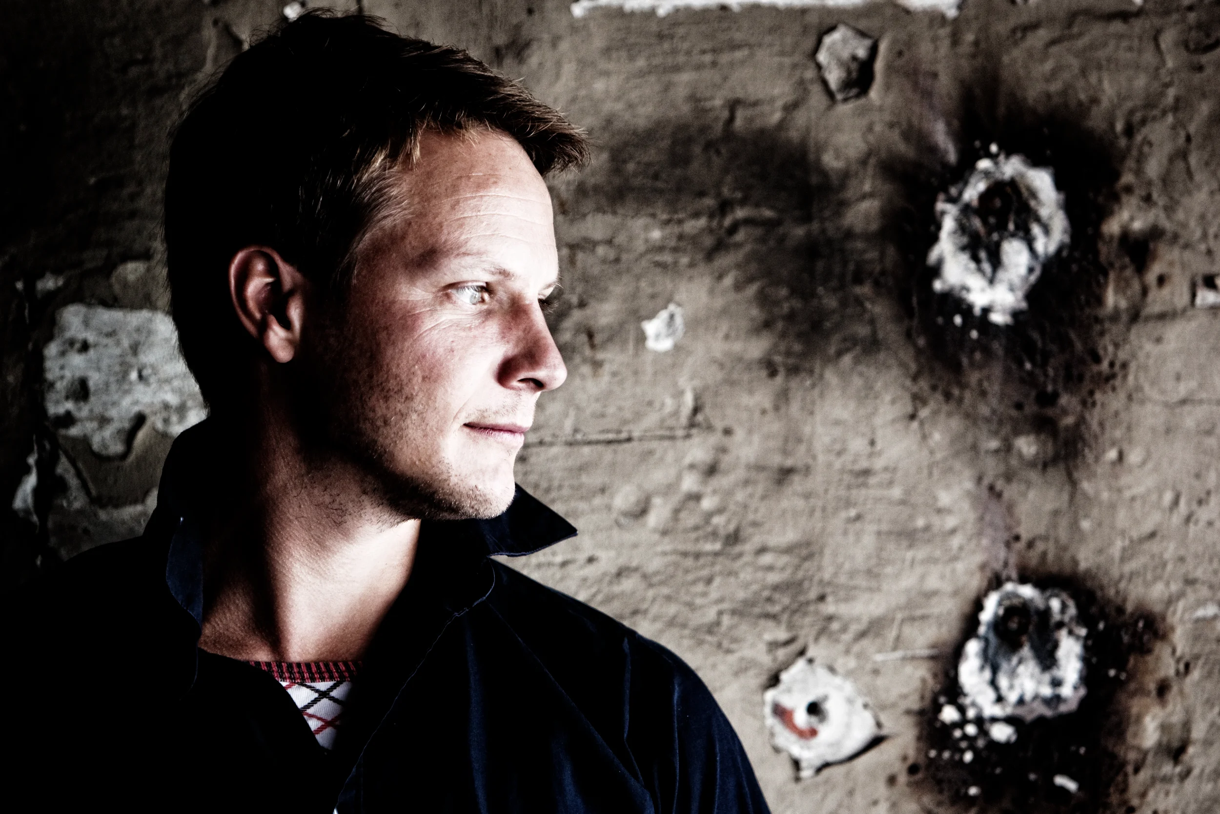 Portrætfoto af mand i profil med intens blik, oplyst af skarpt sidelys mod en rå autentisk baggrund.