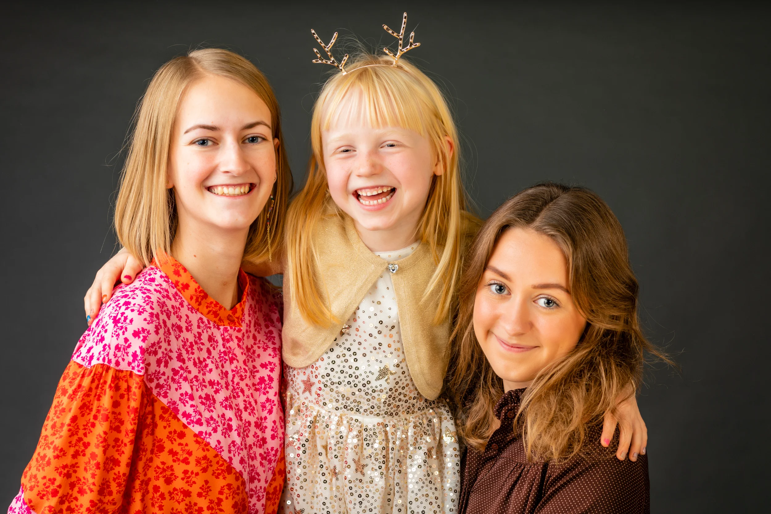 Familiefoto af tre glade søskende mod mørk baggrund, den yngste med et stort smil og gevir-hårbøjle.