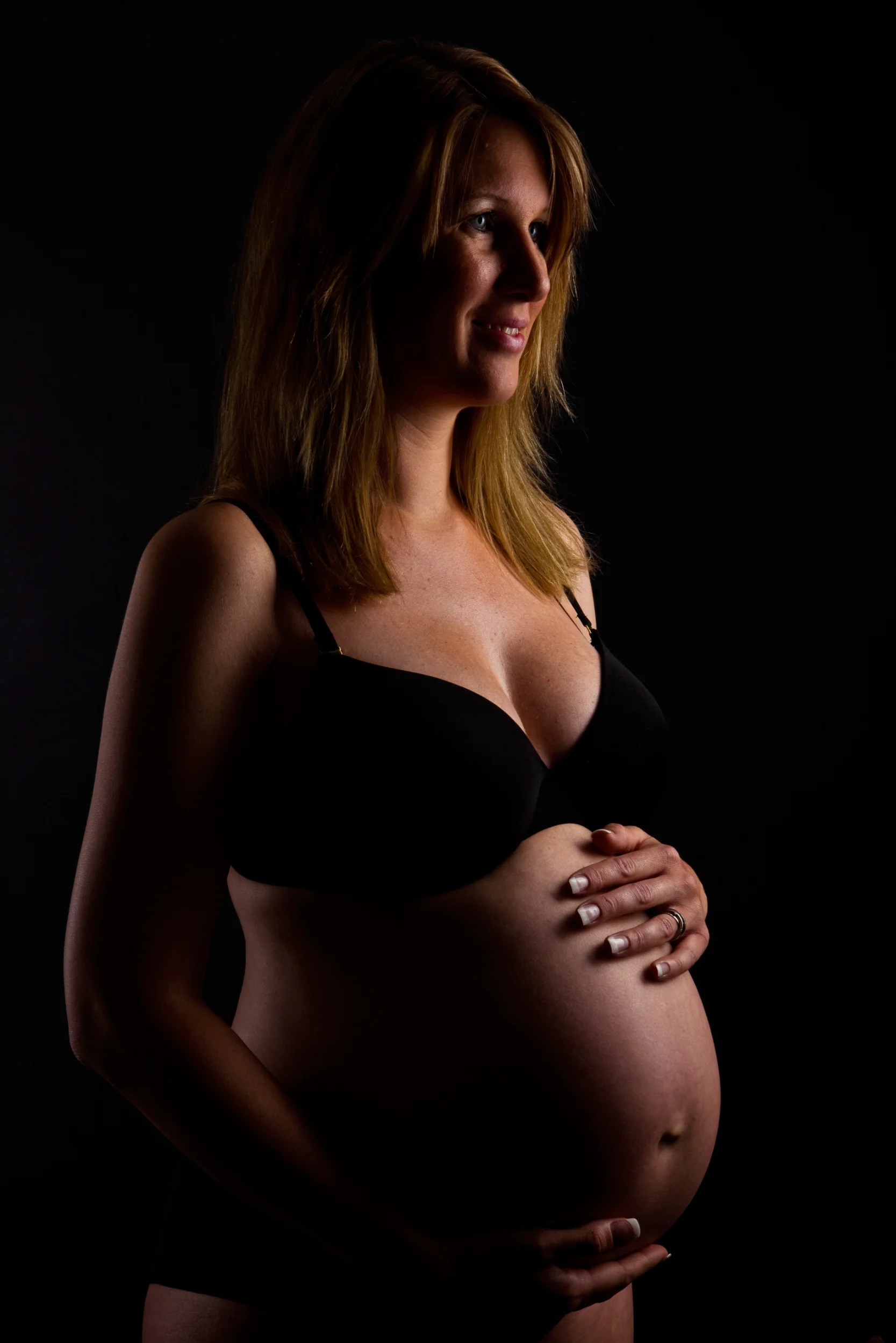 Gravid kvinde i stemningsfuldt lys, der holder sin mave. Et smukt graviditetsportræt i sort/hvid med fokus på bløde skygger.