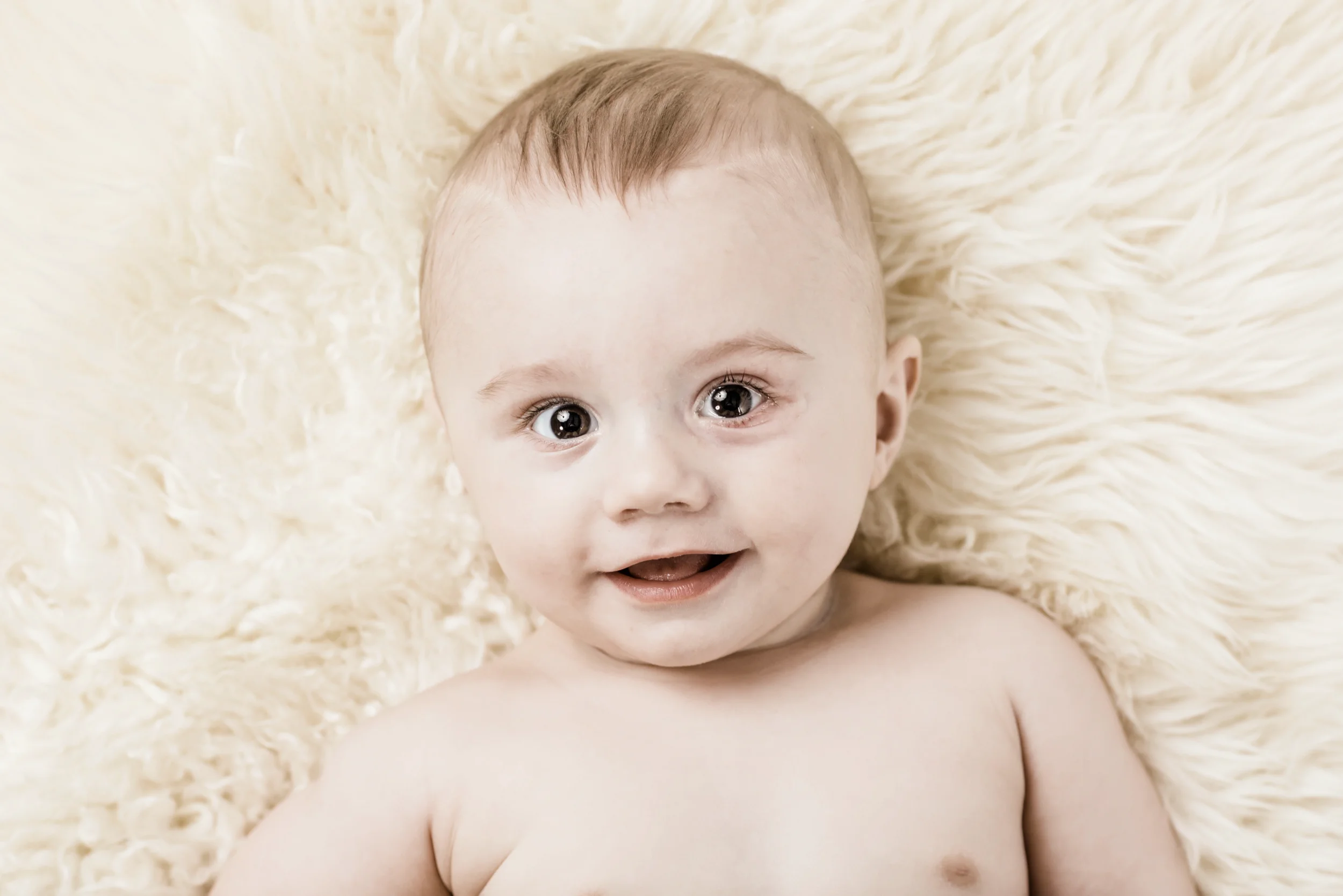Nærbillede af glad, smilende baby med store øjne, der ligger på et lyst, blødt fåreskind. Et naturligt babyportræt.