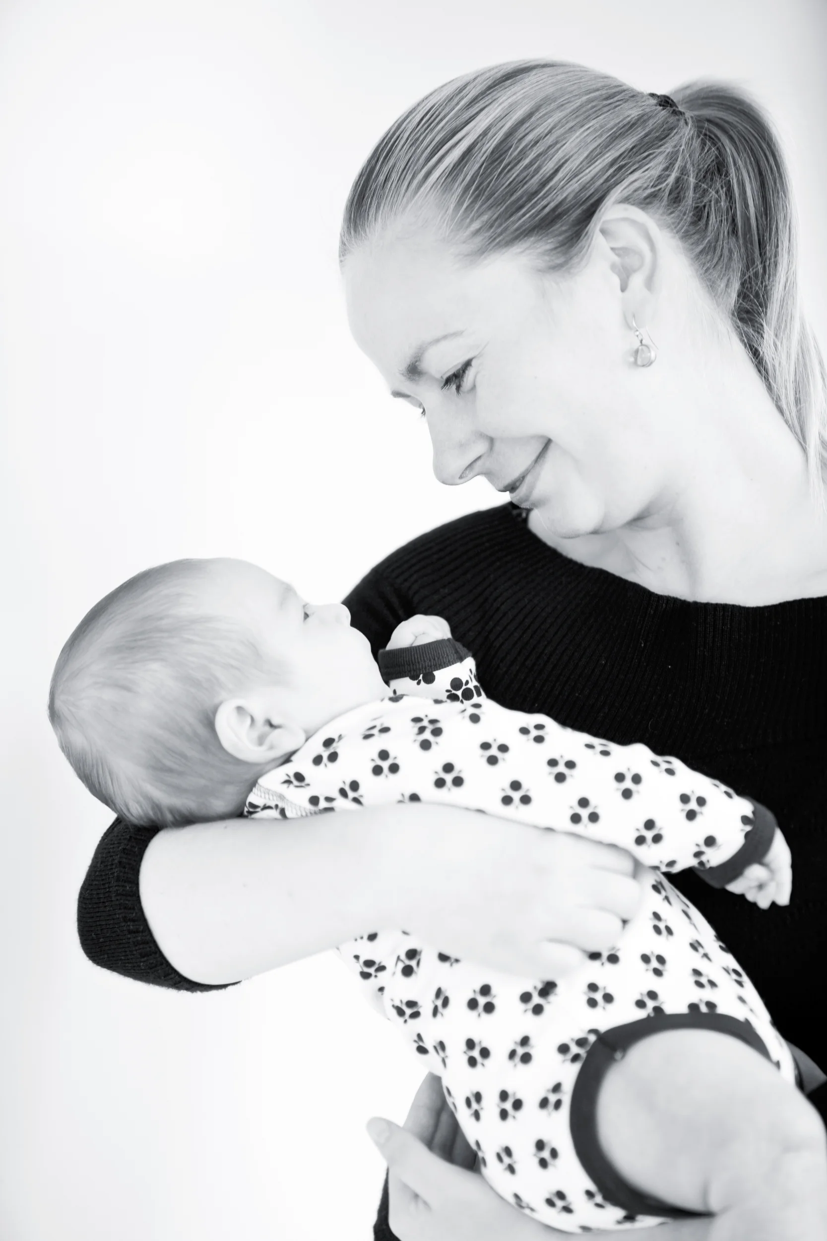 Baby fotografering: Sort-hvidt foto af en mor, der holder sin baby tæt i favnen med øjenkontakt og store smil.