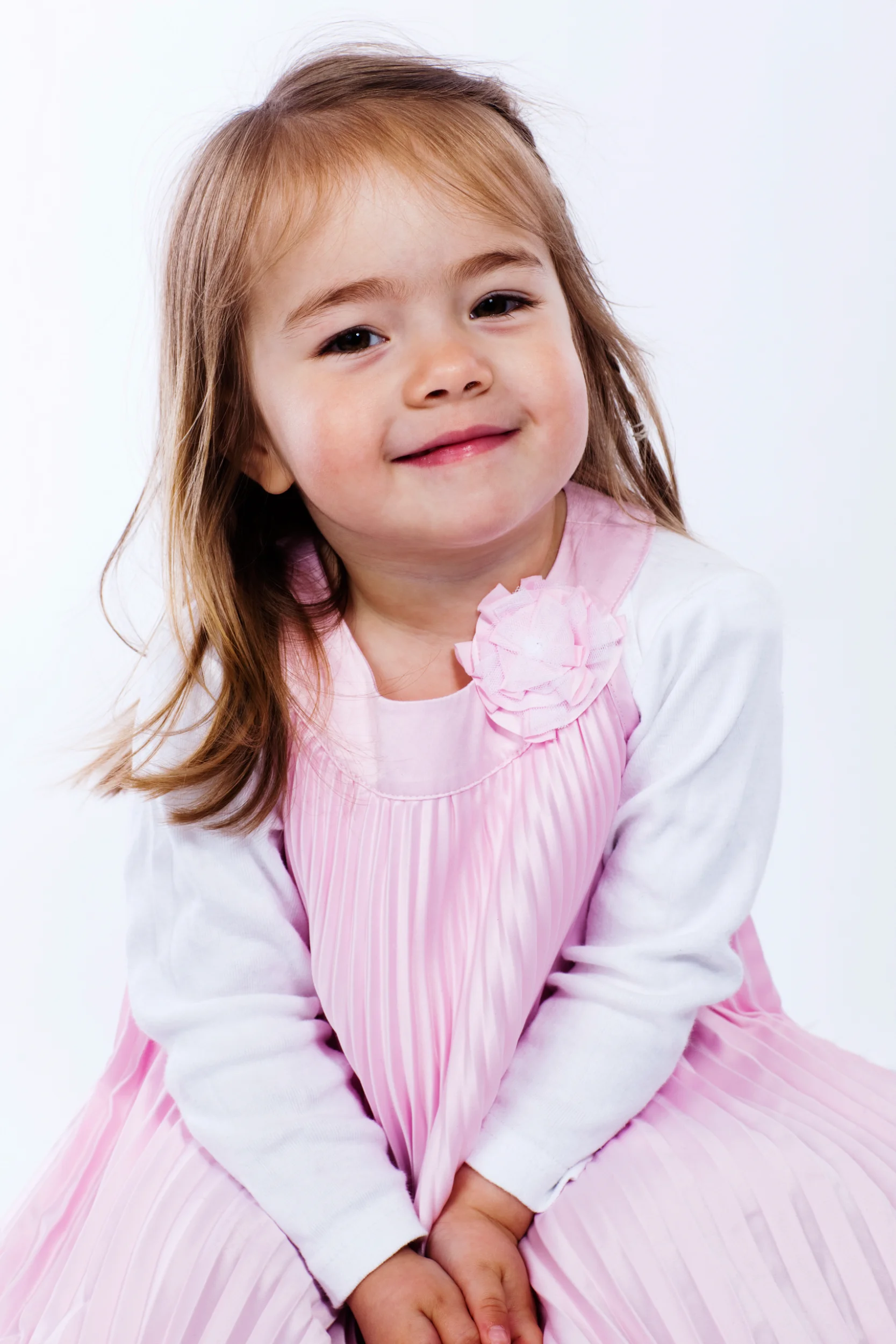 Glad lille pige i lyserød plisseret kjole med blomst på, smilende mod kameraet i et lyst børnefoto.