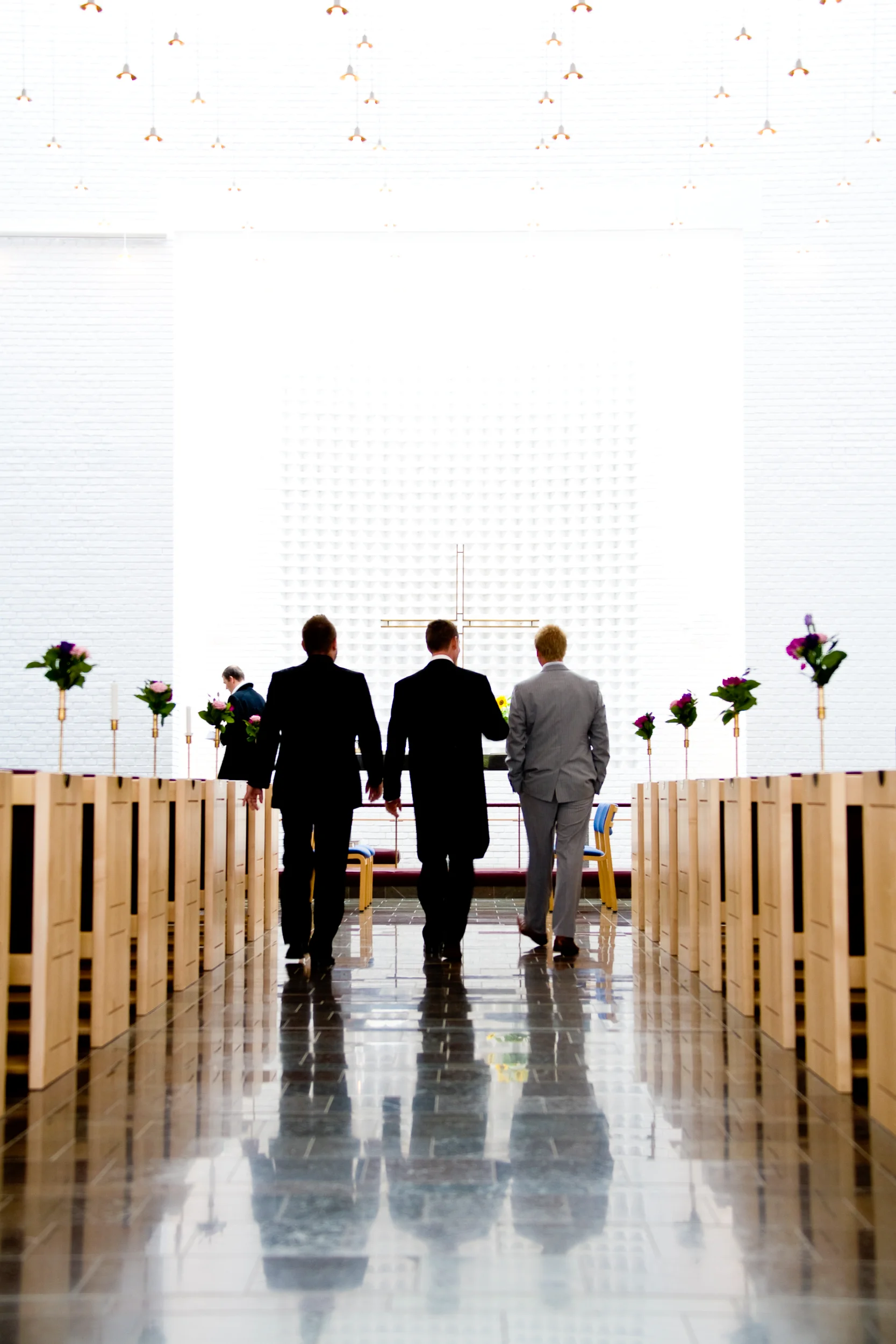 Bryllupsfotografering: Tre mænd i jakkesæt går ned ad kirkegulvet mod et stort kors i et lyst kirkerum.