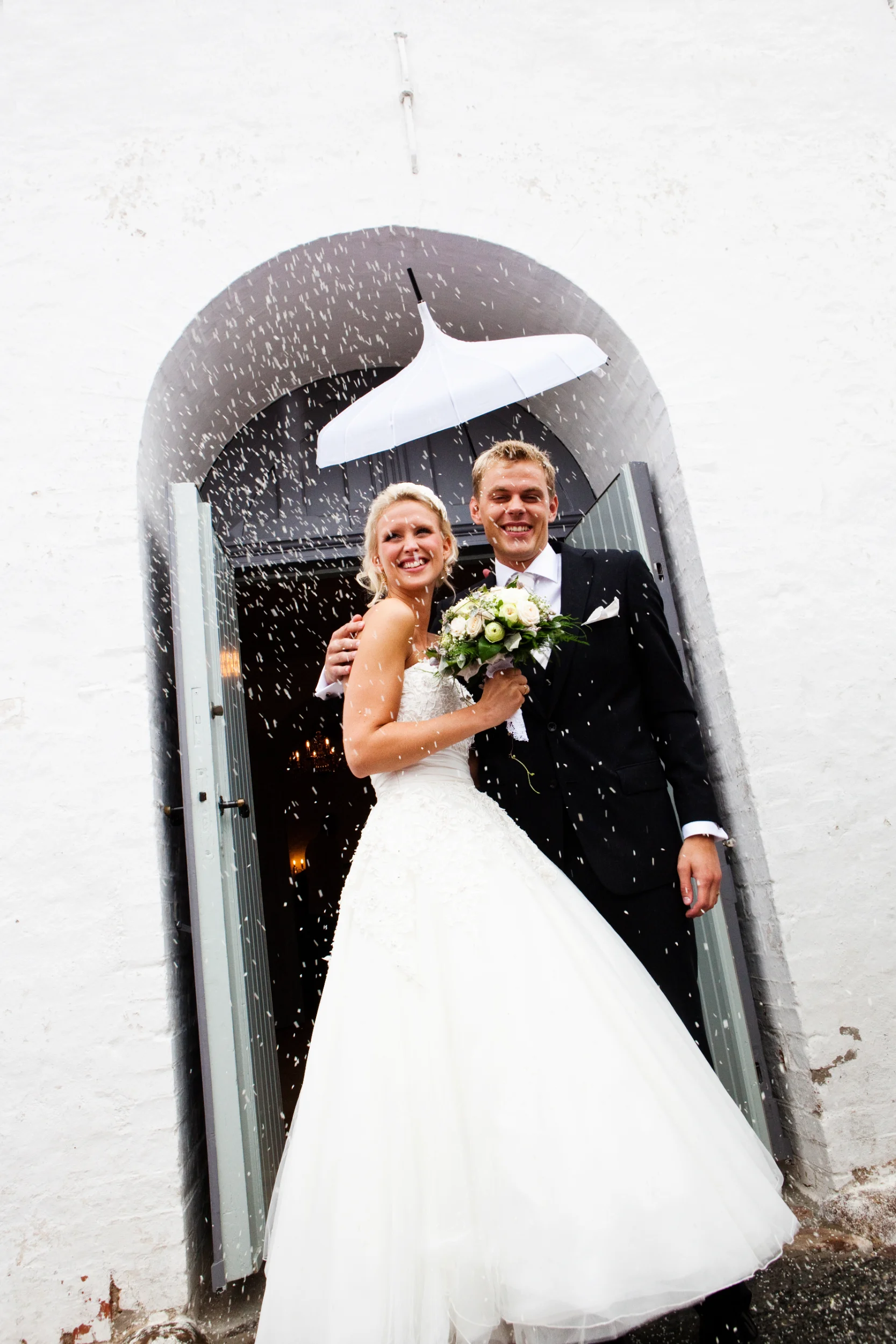 Bryllupsfoto af smilende brudepar i kirkedør med riskast. Bruden i hvid kjole, gommen i sort jakkesæt.