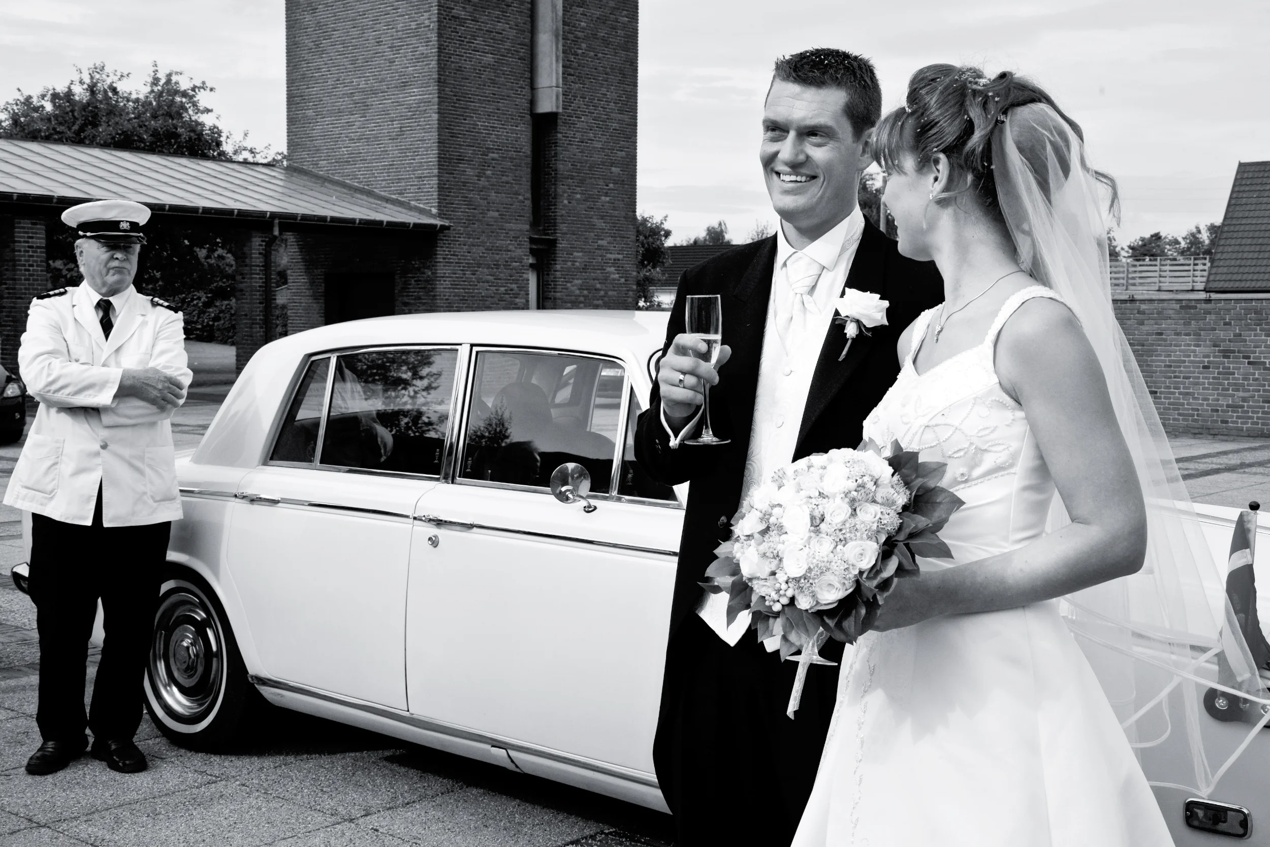 Bryllupsfotografering af smilende brudepar foran hvid veteranbil med chauffør i sort/hvidt billede.