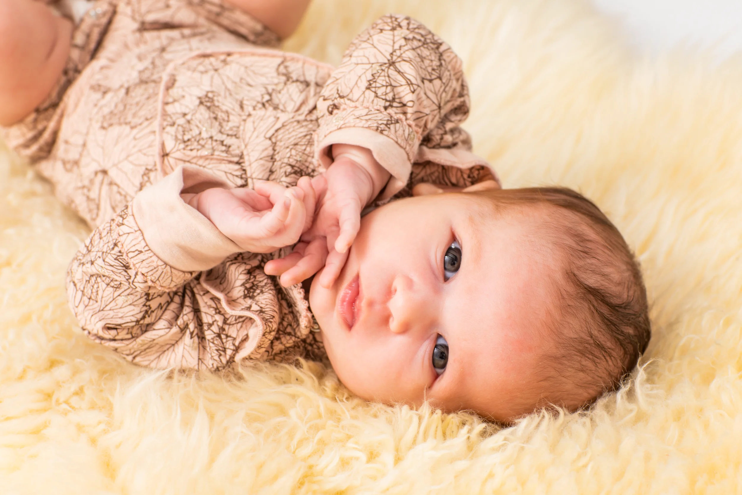 Nærbillede af baby liggende på et blødt, gult lammeskind, iført en dragt med bladmønster. Babyens øjne ser direkte ind i k...