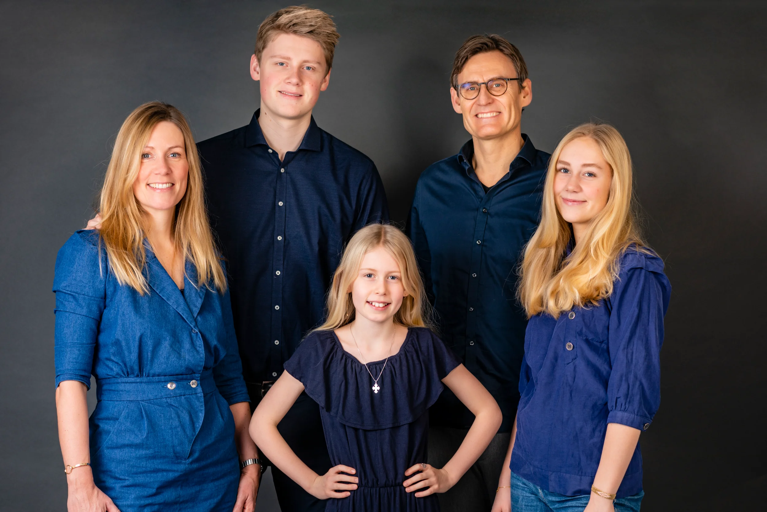 Familiefoto af smilende mor, far og tre børn i blå nuancer mod mørk baggrund. Glade ansigter, klassisk børnefotografering.