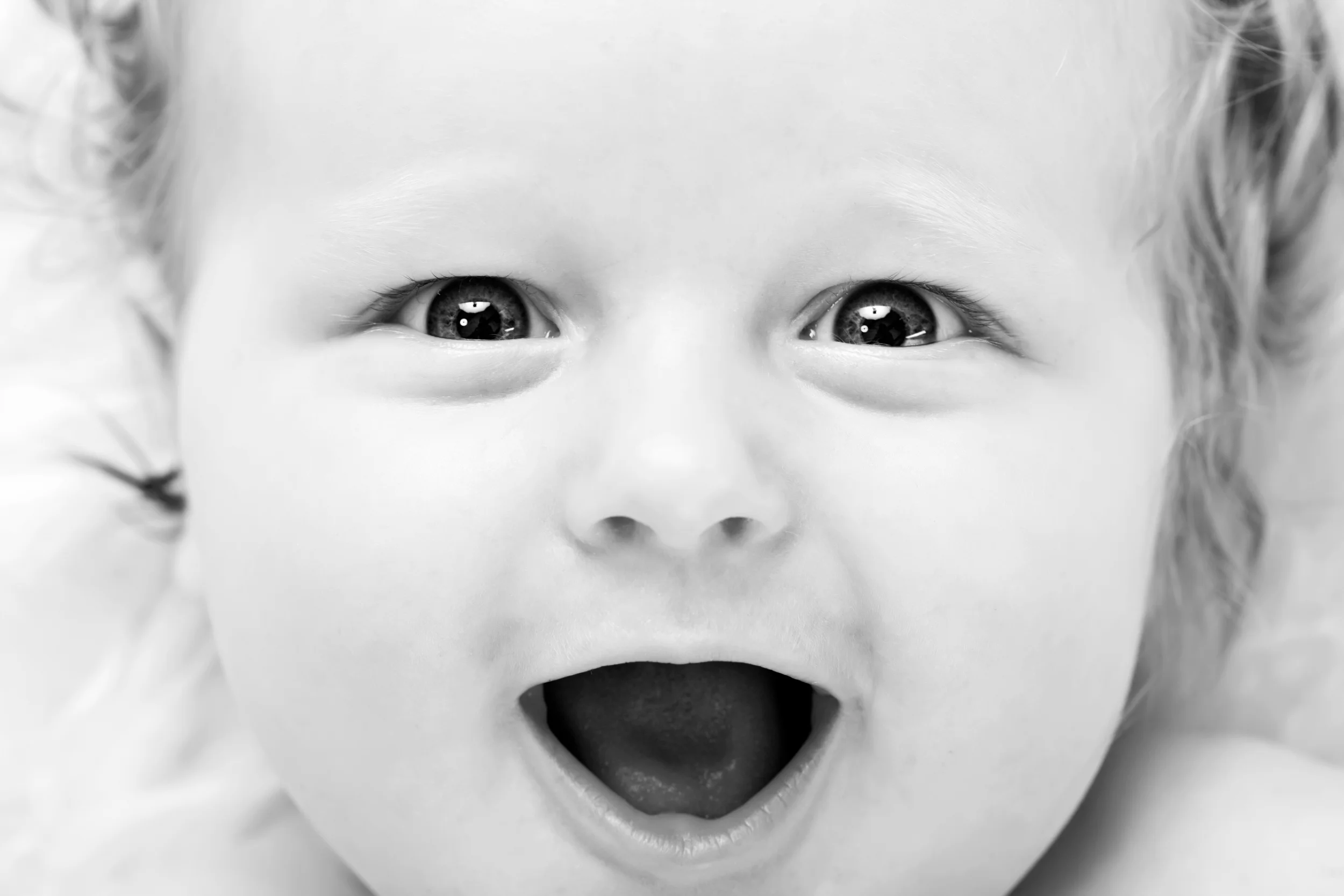 Nært babyfoto i sort/hvid af nysgerrigt ansigt med åben mund og store øjne, der fanger opm lyset. Skarpt og sødt. Fra en f...
