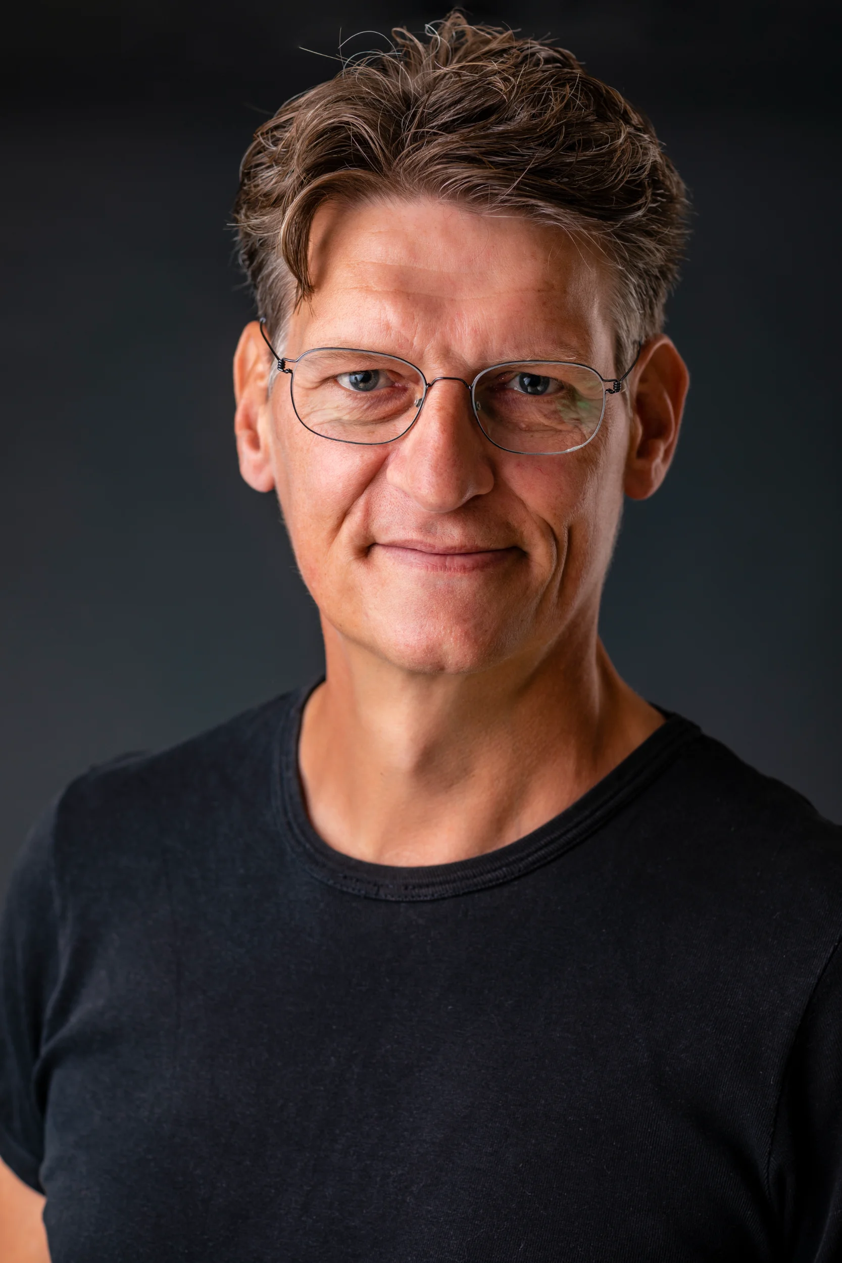 Portrætfoto af mand med mørkt hår og briller, smilende mod kameraet på en mørk baggrund.