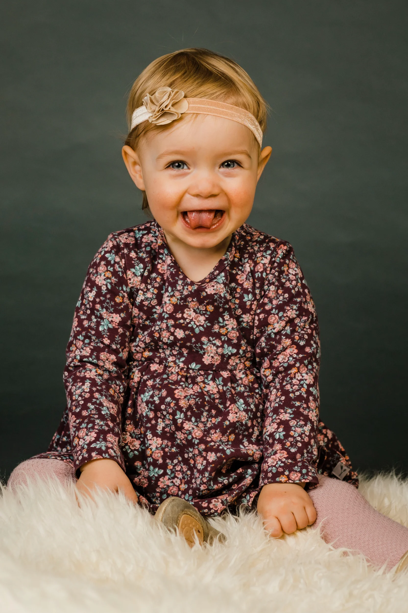 Glad barn i blomstret kjole og pandebånd sidder på hvidt lammeskind, rækker tunge mod kameraet i Virum.