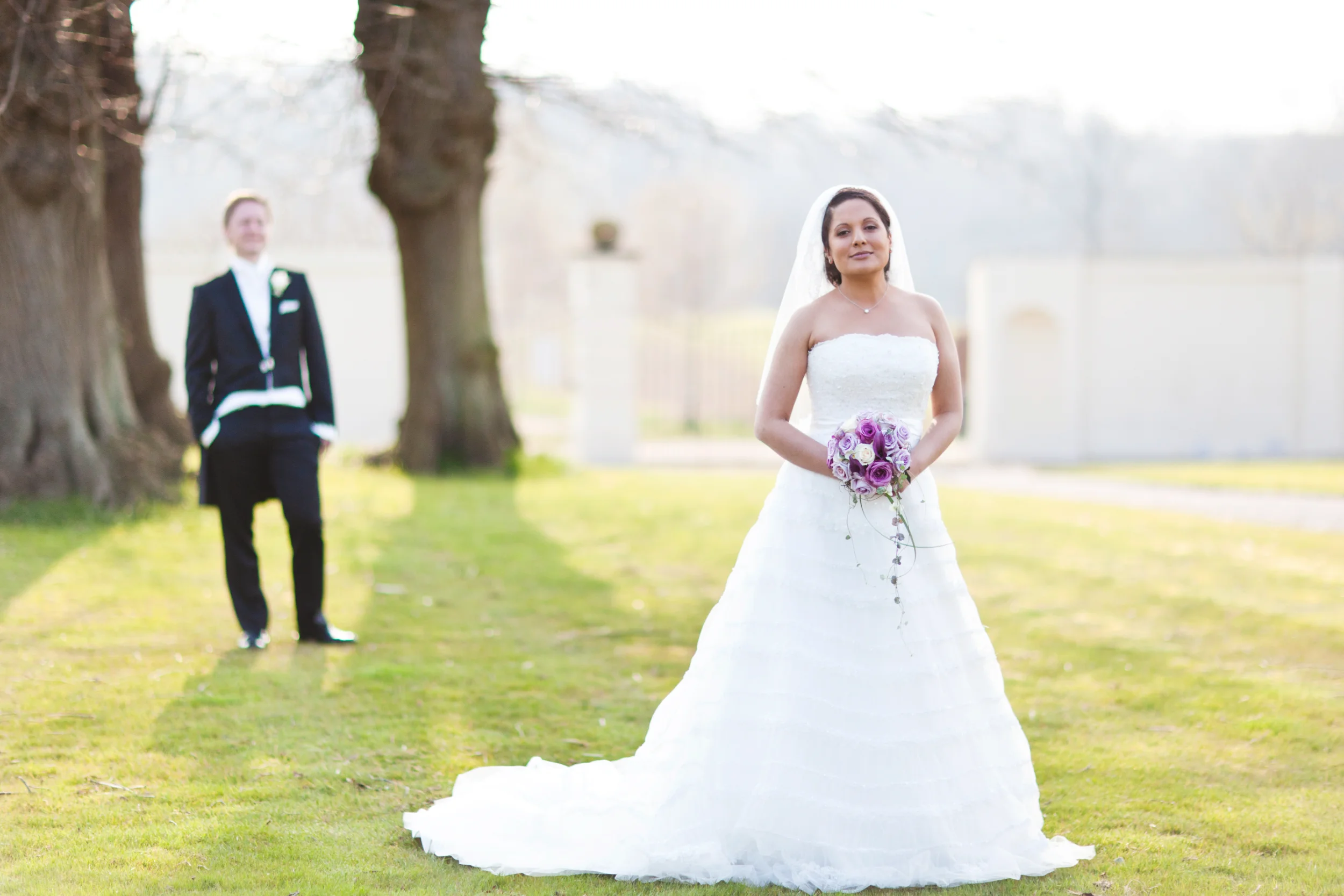 Bryllupsfotografering i sort/hvid af en brud i hvid kjole med brudebuket, stående på en græsplæne. Brudgom i baggrunden.