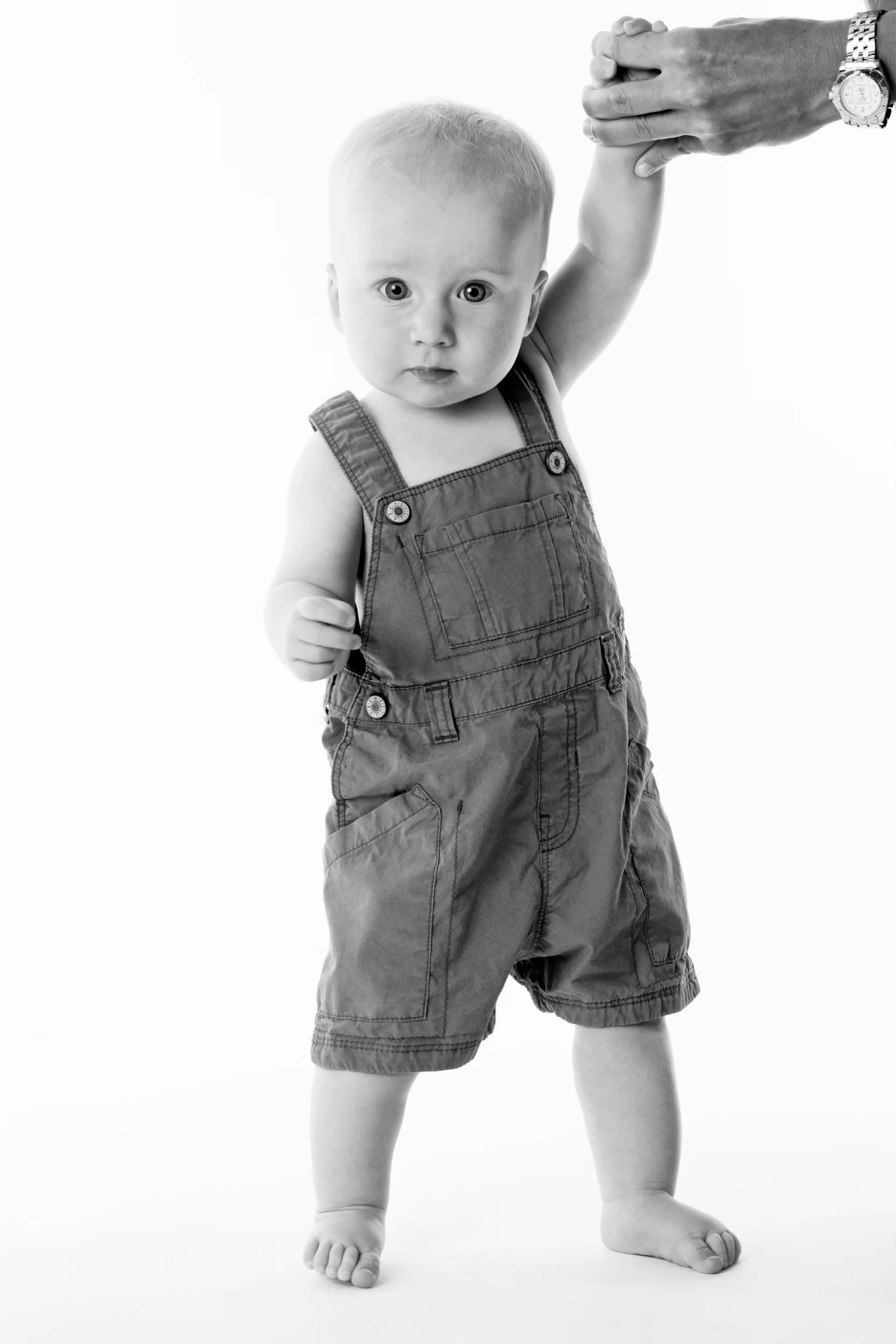 Baby i blå selebukser, der holder en hånd for at balancere, med nysgerrigt blik mod kameraet.